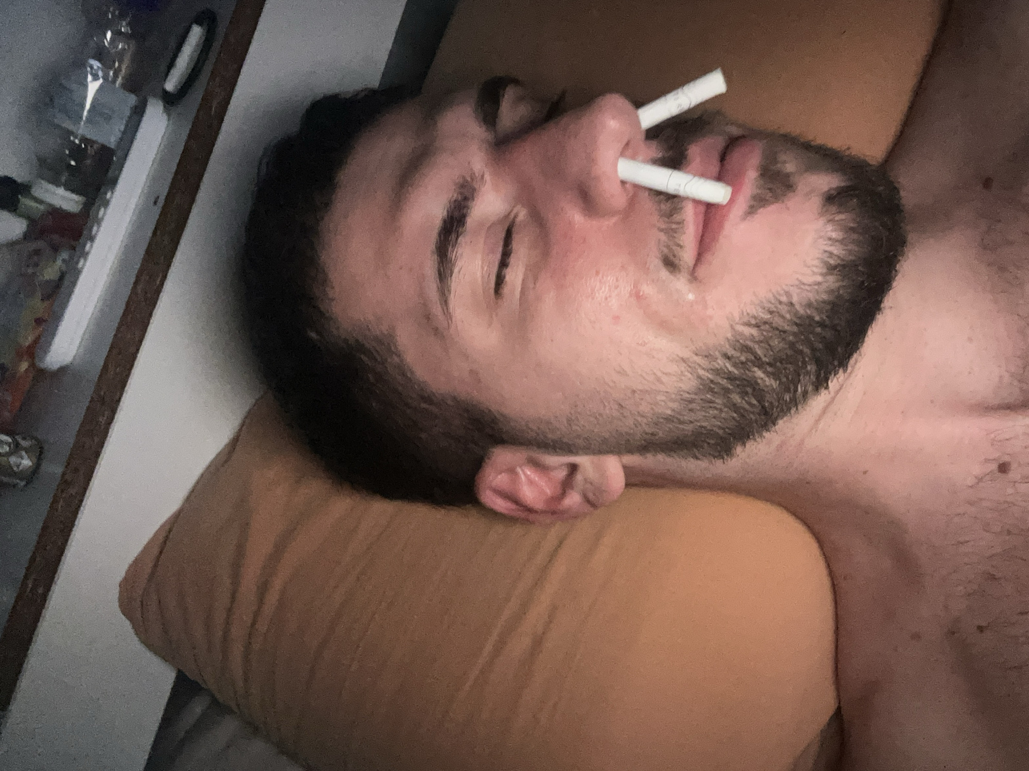 A medium-shot depicts a male lying on a bed, resting on an orange pillow. Two white cigarettes are stuck in his right nostril and mouth. His eyes are closed. He has dark hair and a beard. A section of a shelf containing an indiscernible collection of items is visible on the left. The location is not discernible from the visual content provided. No city-specific details regarding Sofia, Bulgaria, are present.