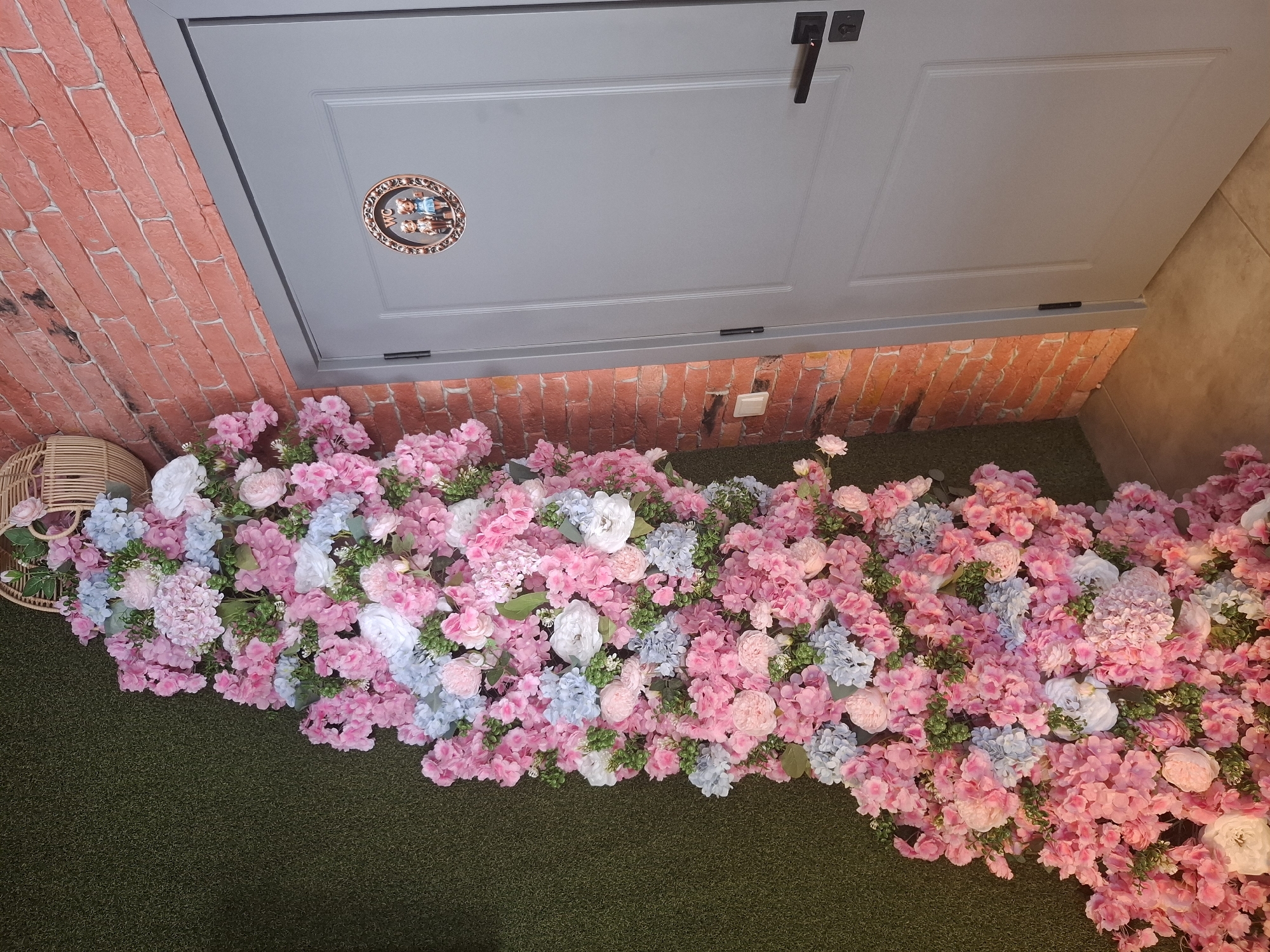 The image depicts a decorative floral arrangement featuring pink, white, and light blue flowers. The arrangement is positioned vertically against a wall covered in artificial green turf on the left side of the frame. To the right of the floral display is a grey door with a black handle and a small circular plaque above it. This plaque contains a decorative border and features the letters "WC" along with two small figures, suggesting it is a restroom door. The wall to the right of the door is constructed of red brick. A light switch is visible on the brick wall near the bottom left of the door. The floor appears to be tiled. The location is identified as Dubăsarii Vechi, Moldova. There are no people visible in the image.