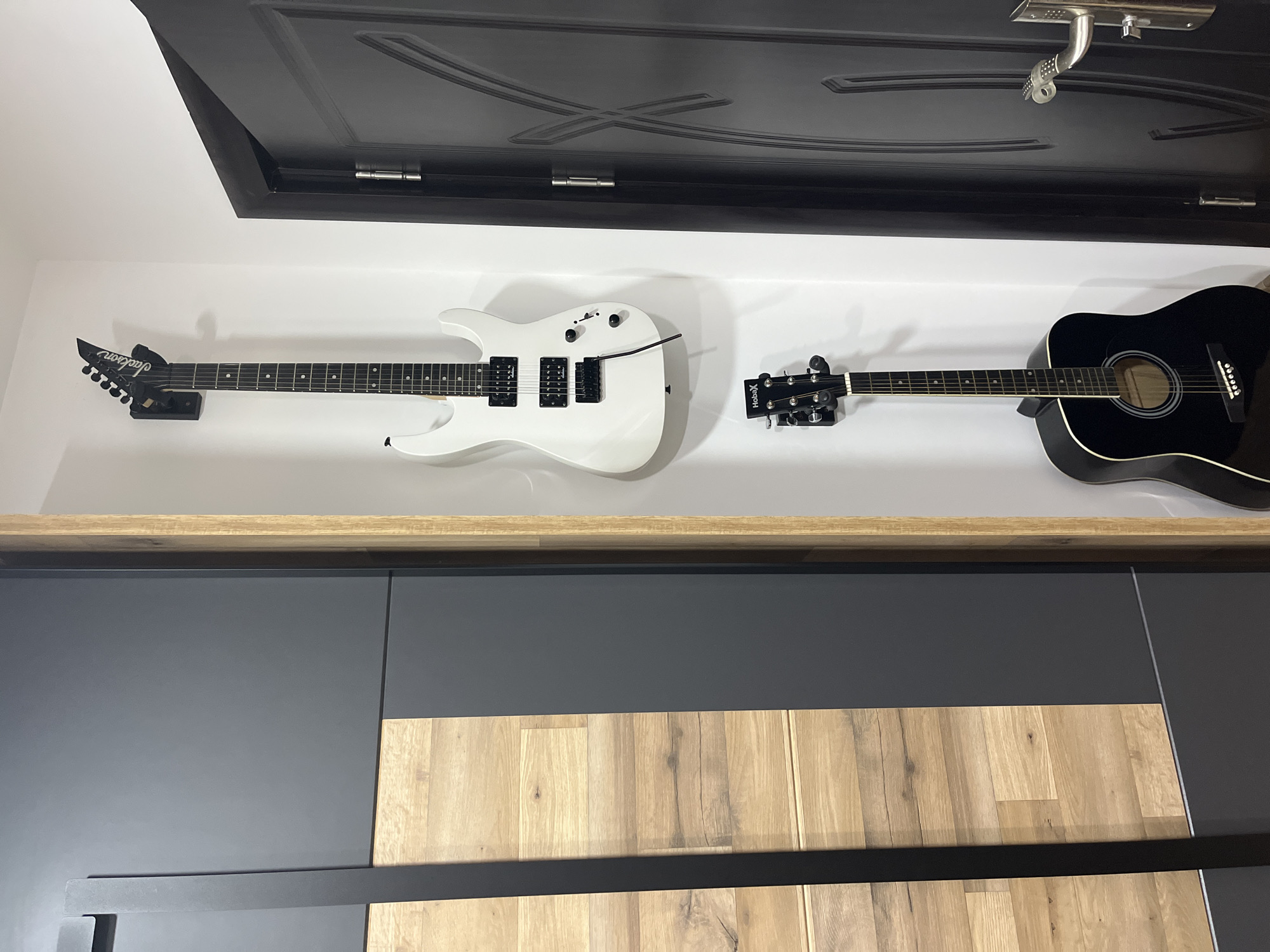 Here is a detailed description of the image:
The image presents a close-up of an interior space, featuring two guitars mounted on a wall above a shelf. The wall is painted white, and a dark, possibly black, door frame is positioned at the top of the frame. The guitars include a white electric guitar on the left with a black headstock and black hardware. To the right is a black acoustic guitar. Both guitars are hanging on wall mounts.
Beneath the guitars, a wooden shelf extends across the width of the frame. Below the shelf are dark gray cabinets with vertical wood panels. The combination of the wood and dark gray gives the cabinets a modern and somewhat contrasting design. A black metal handle runs horizontally across the cabinets. The lighting in the room appears soft and diffused.
The scene does not provide enough information to identify a specific location or city.