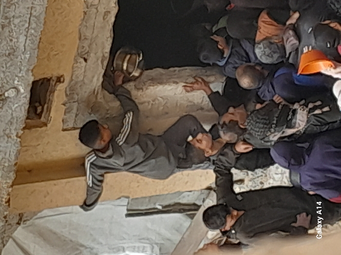 In Khan Yunis, Palestinian Territory, a crowd of individuals is gathered within what appears to be a partially damaged or makeshift interior space. A young male, wearing a grey tracksuit with white stripes, stands elevated on another person's shoulders. He extends his right arm into an overhead aperture in a rough, possibly concrete, wall, holding a metallic bowl. His left hand is pressed against the wall for balance. The person supporting him, also a young male in a dark shirt, looks upwards with his right hand partially raised. Below them, a dense group of individuals, predominantly males, observe the activity. Their attire consists of various dark-colored clothing, including a patterned headscarf on one individual, and an orange hard hat brim is visible in the lower right corner. The walls and ceiling exhibit unfinished or damaged construction materials.