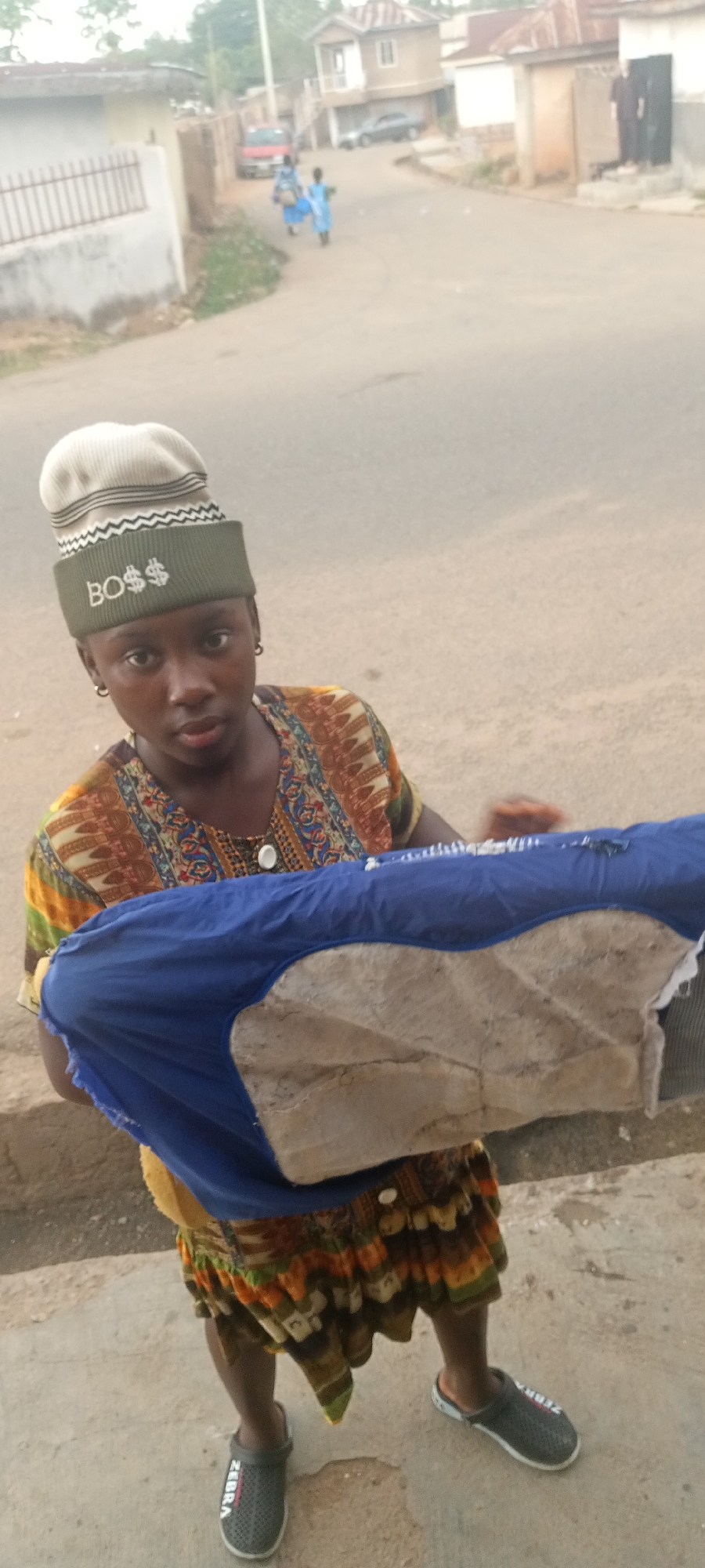 This media file captures a street scene in Jos, Nigeria, primarily focusing on a young person in the foreground. The individual, appearing to be female with dark skin, looks directly at the camera with a neutral expression. She wears a distinctive light beige and olive green knitted beanie hat adorned with a zig-zag pattern and the stylized text "BO$$" (with dollar signs replacing the 'S's). Her attire includes a vibrant, multi-colored patterned short-sleeved top featuring earthy tones of brown, orange, green, and blue, along with a matching patterned skirt. On her feet are dark grey clogs with white soles, one of which clearly displays the brand name "ZEBRA".
The person holds a large, bulky item wrapped in blue fabric, which appears to conceal a lighter, possibly cardboard or sack-like material, suggesting she might be transporting or selling goods.
In the blurred background, a paved road winds through a residential area. Two smaller figures, likely children dressed in light blue clothing, walk away from the camera further down the street. Houses with varied architecture, including single and multi-story structures with fences, line the road. A red car is parked on the left side, with another vehicle visible in the distance. The ambient lighting suggests it is daytime, possibly late morning or afternoon, with no harsh shadows, indicating fair weather conditions. The overall setting depicts a typical urban or semi-urban environment in Nigeria.