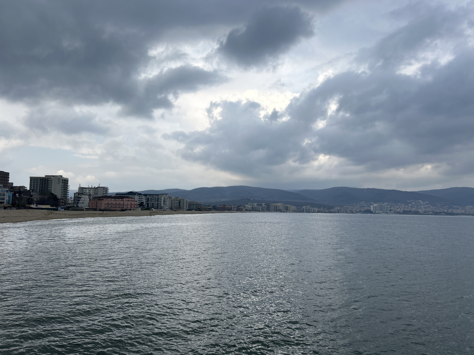 The image shows a seascape under a cloudy sky. In the middle ground, there is a body of water with visible ripples on its surface. Along the shore in the distance, a line of buildings suggests a coastal city, with mountains visible in the background. The light is subdued due to the overcast conditions. No people are clearly discernible in the image.