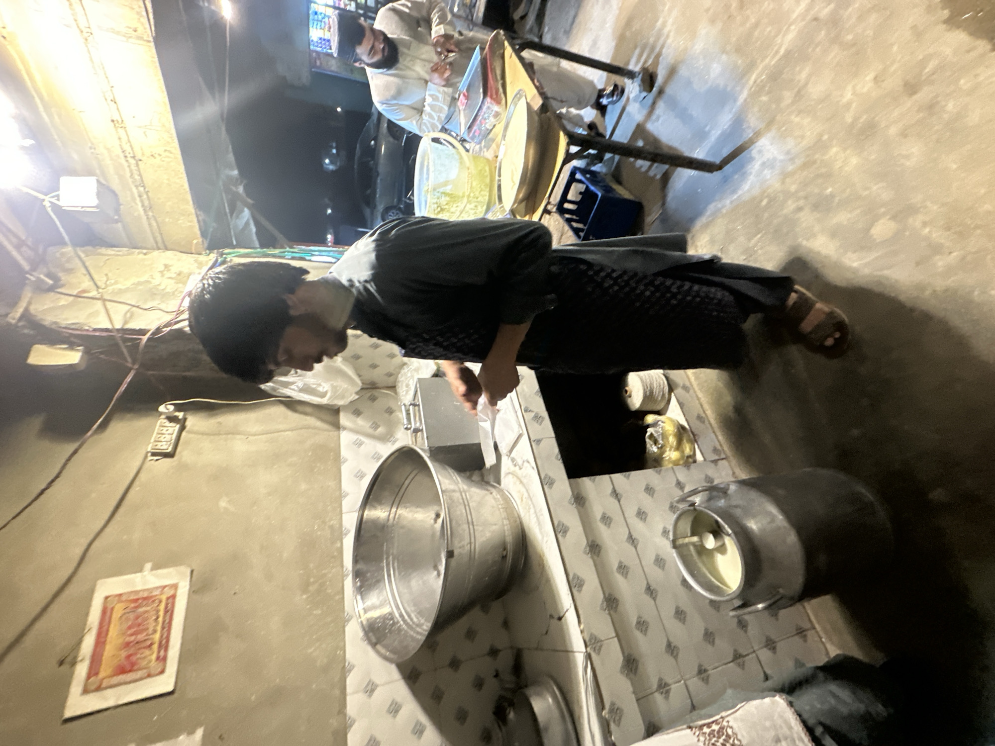 The image shows an outdoor food preparation area in Quetta City Tehsil, Pakistan. A person wearing a dark garment and sandals is seen near a tiled counter. On the counter are a large metal bucket, a small metal container, and other items. In the background, another individual in a white garment is visible, seemingly serving customers from a food stall. Overhead lighting is present. The environment is urban, with electrical wiring and a framed print on a wall nearby.