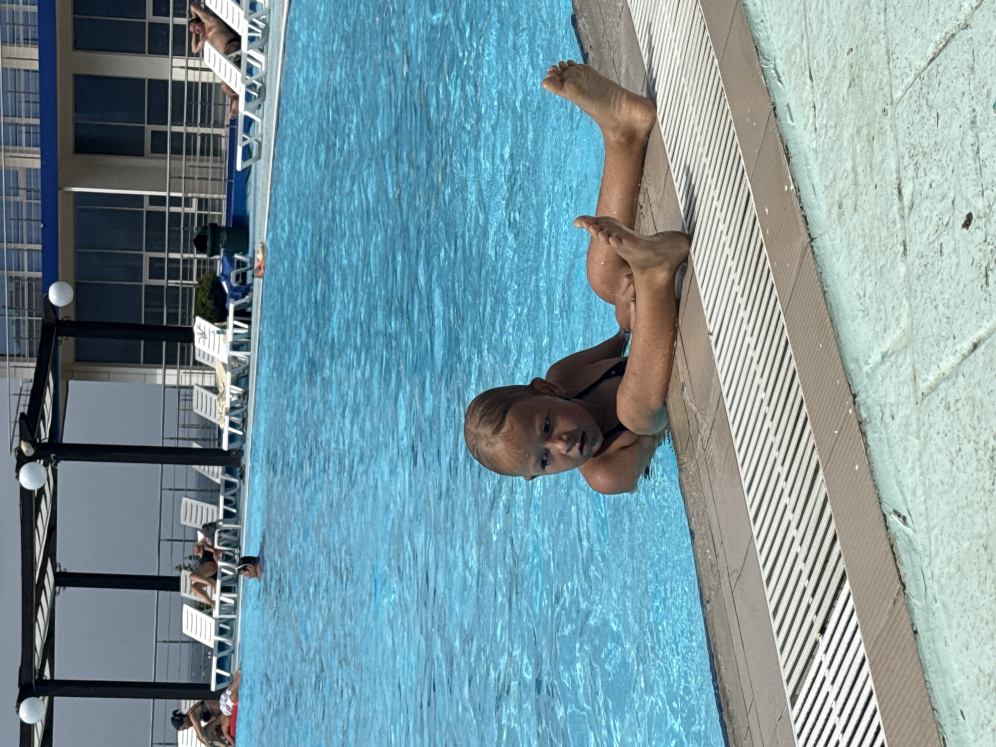 The image depicts a young girl with blonde hair, wearing a black swimsuit, hanging onto the edge of a swimming pool. Her legs and feet are visible, bent at the knees and resting against the pool's edge. She is looking towards the camera with a curious expression. The water in the pool is a clear, vibrant blue, reflecting the sunlight. The pool area appears to be outdoors, with lounge chairs and a building visible in the background. The time of day seems to be sunny, likely daytime, given the brightness and the clear blue water. The scene is set at a swimming pool, possibly a resort or a public facility, with the architecture of the building suggesting a warm climate. There is no visible text in the image.