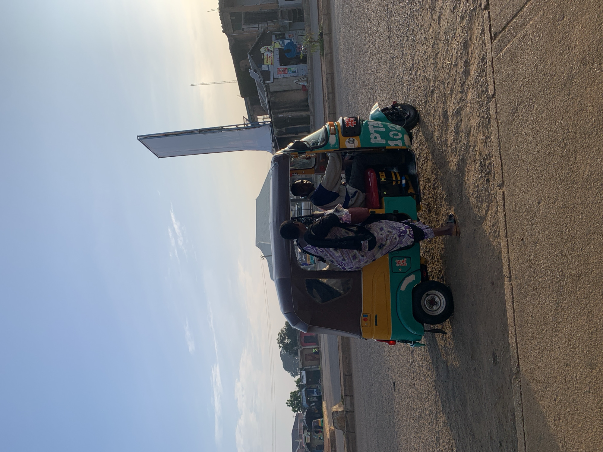 A man drives a motorized tricycle, painted yellow and teal with "PTA 102" in blue lettering on the side, down a dusty road in Rafin Par, Nigeria. A woman wearing a purple and white patterned garment stands beside the tricycle, appearing to be interacting with the driver. In the background, several modest buildings line the street, and a tall, narrow billboard stands in the distance against a clear blue sky. The ground is a mix of dirt and asphalt, with shadows cast by the tricycle and its occupants. The scene is illuminated by natural light, suggesting it is either early morning or late afternoon.