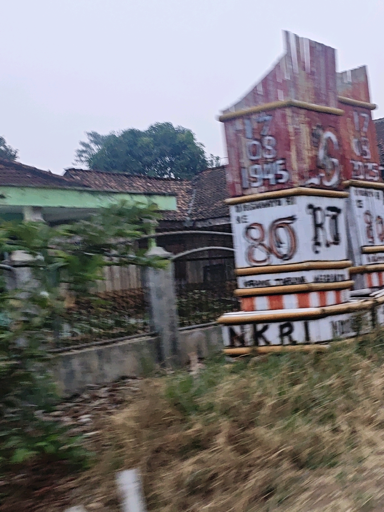 The image depicts a somewhat blurry, outdoor scene in what is identified as Karang Sari, Indonesia. The focal point is a weathered, red-and-white monument with bold lettering and numbers. The top portion of the monument features what appears to be a stylized flag or banner, painted in streaks of red. Below this, large numbers are painted, including "17 03 1945" and "17 03 2015", suggesting historical significance, possibly related to Indonesian independence.
Further down the monument, more text is visible, with a prominent "80" and "RI" painted in a graffiti-like style. Below this, a band of red and white stripes runs horizontally, bordered by yellow. The bottom section of the monument displays the letters "NKRI", which stands for Negara Kesatuan Republik Indonesia (Republic of Indonesia).
The monument is situated in a slightly overgrown area with dry grass and foliage in the foreground. In the background, traditional Indonesian houses with brown tiled roofs and green trim are visible. Trees with lush green foliage form a backdrop behind the houses. The overall lighting suggests an overcast or early morning/late afternoon sky, contributing to the slightly muted colors and soft focus of the image. There are no people visible in the scene. The motion blur suggests the photograph was taken from a moving vehicle.