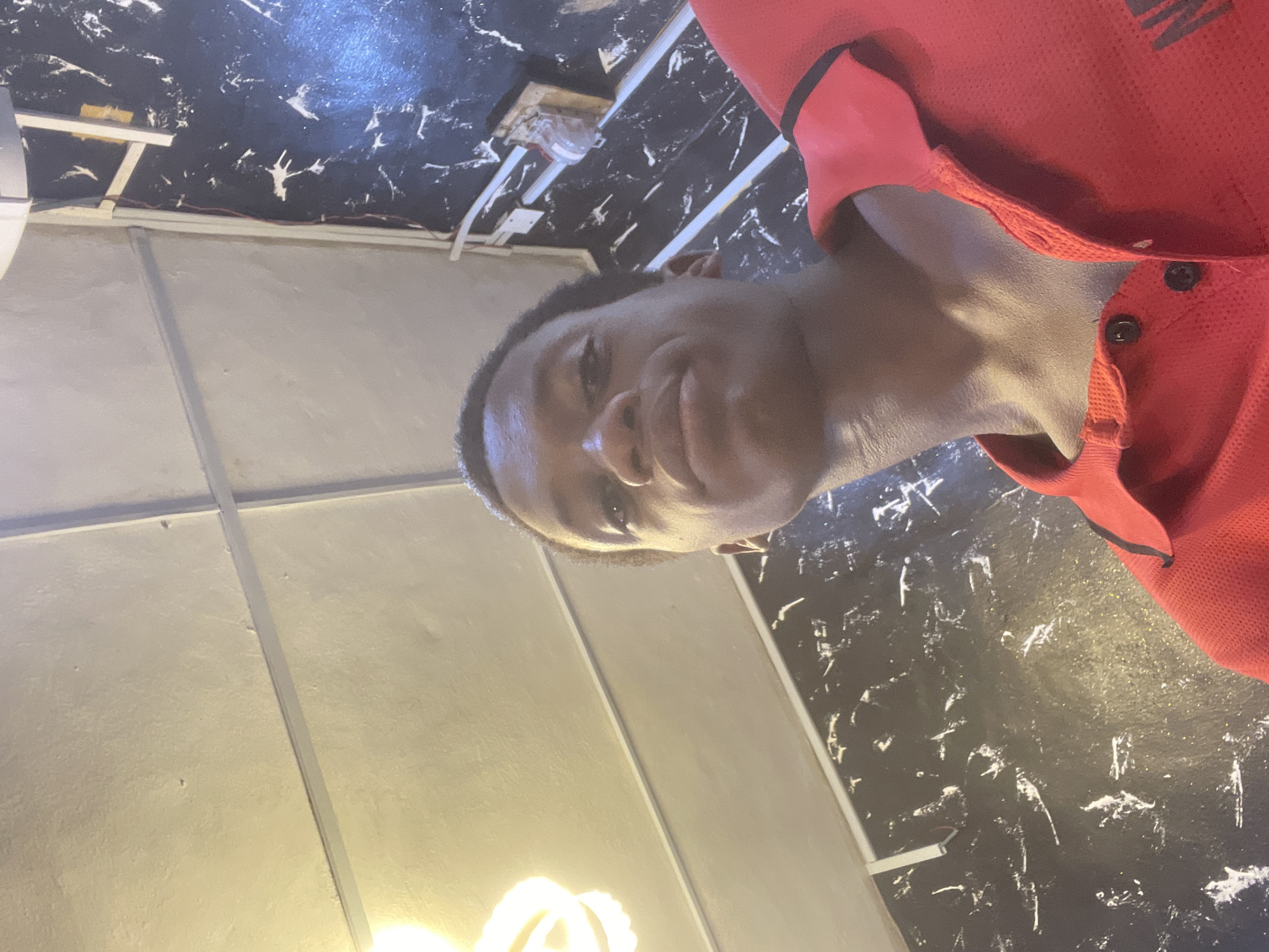 A young man, likely of African descent, is the central subject of the close-up, informal portrait, captured in what appears to be a selfie. He looks directly at the camera with a neutral to slightly relaxed, pouting expression. He has short, dark hair and is wearing a red polo shirt with a textured, possibly athletic, fabric, featuring a collar and black buttons.
The background reveals an indoor setting. To the man's right, a dark surface, possibly a wall or ceiling, is decorated with abstract white, paint-splatter-like patterns. Visible on this dark surface are electrical wires and a small fixture or outlet. To the man's left, a lighter, off-white or light grey ceiling section with structural beams or panels is present. A bright, warm light source, potentially an exposed light bulb, emanates from the lower-left portion of the background, casting a soft glow.
The image conveys a casual moment indoors, possibly in a room or commercial space in Onitsha, Nigeria. No specific activity beyond posing for the picture is evident. There is no visible text within the image.