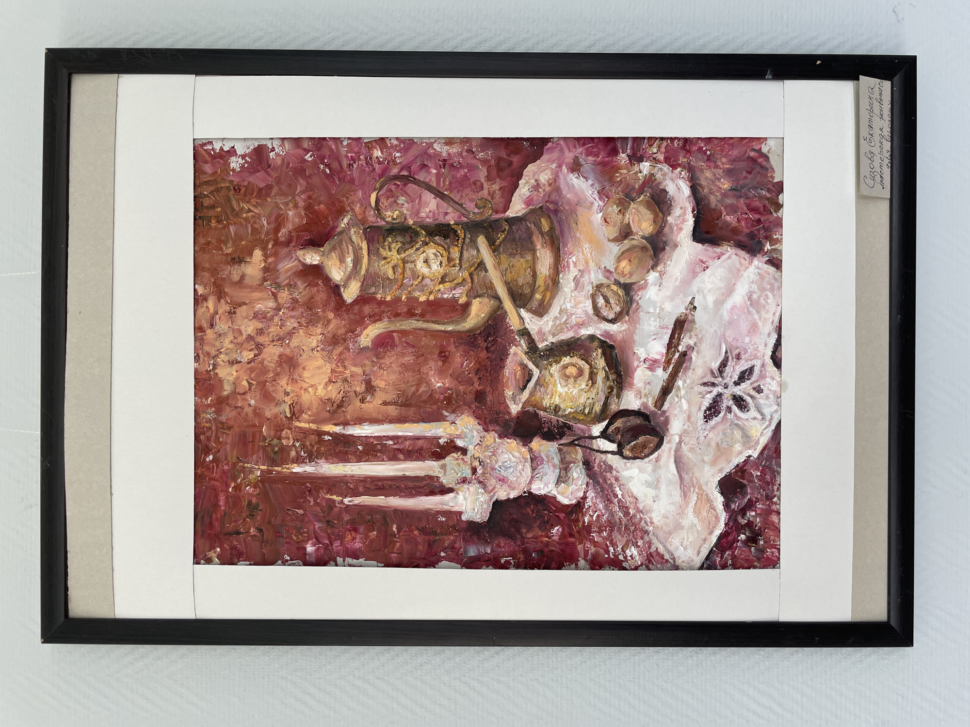 This is a photograph of a framed painting. The painting is a still life featuring a golden, ornate coffee maker with a long spout and handle, positioned on a textured, deep red background. To the right of the coffee maker, a white cloth is draped, revealing several nuts and two small dark objects that appear to be coffee beans or chocolates. The cloth has a floral pattern embroidered in a darker shade of purple or brown. In the foreground, on the left side, are three white objects resembling cloves or perhaps stylized flowers, arranged horizontally. On the right side, near the cloth, are what appear to be scissors. The painting style is impasto, with thick, visible brushstrokes, particularly in the textured background and the white cloth. The colors are rich and warm, dominated by reds, golds, and whites, with touches of brown and purple.
Attached to the right side of the frame is a small, light-colored label with handwritten text in Cyrillic script. The text appears to read "Сазонова Екатерина / Диадема Растение / Алая Сирень", which translates to "Sazonova Ekaterina / Diadema Plant / Scarlet Lilac". The painting is mounted within a black frame, with a double mat: an inner white mat and an outer cream-colored mat. The overall presentation suggests a finished artwork ready for display. The lighting in the photograph is even, illuminating the painting without harsh shadows, and the background is a plain, light-colored wall. No specific time of day, weather conditions, or explicit emotional cues are discernible from the painting itself or its presentation. The location context is given as Perm, Russia.