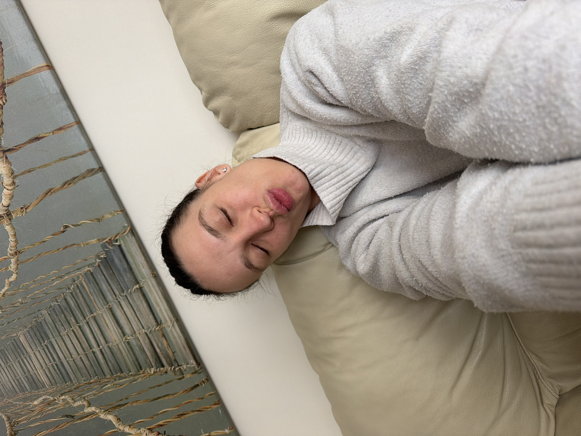 A woman lies on a light-colored leather couch, wearing a light gray sweater. Her eyes are closed and her lips are pursed. An off-white pillow is behind her head. To the left of the couch is a piece of art depicting what appears to be a bridge. The location is likely an indoor setting, possibly a residence or hotel, but the city of Budva, Montenegro, cannot be determined from the image.