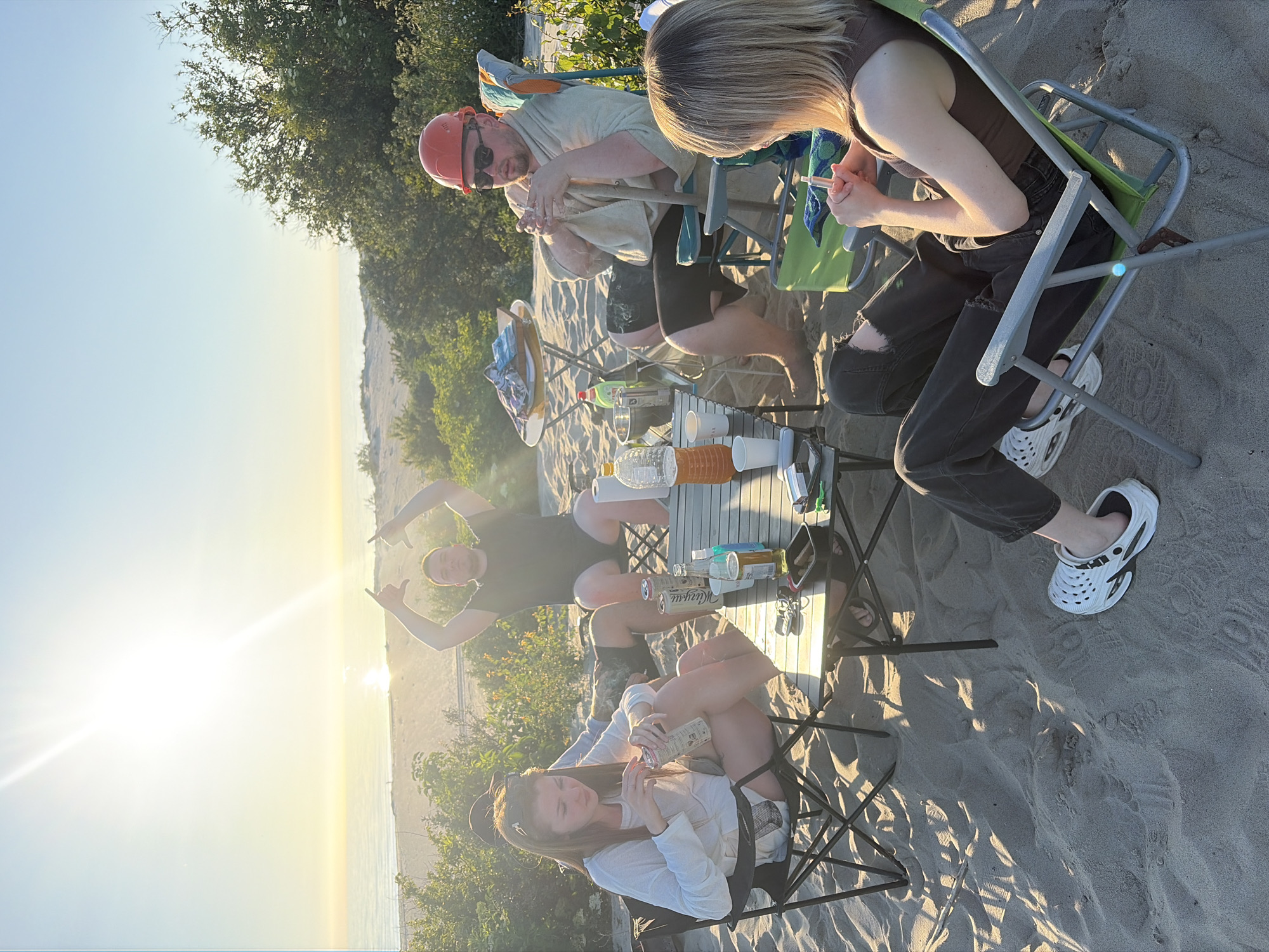 Five people are gathered on a sandy beach, likely for a picnic or small social gathering. They are seated in camping chairs around a low-lying table laden with drinks and snacks. A man in an orange hard hat and sunglasses is in the foreground, holding a cigarette. To his right, a woman with blonde hair is seated, also holding a cigarette. Two other individuals are visible in the background, one with their arms raised in a "shaka" gesture. The sun is low in the sky, casting a warm glow and long shadows across the sand, suggesting it is either sunrise or sunset. The presence of trees and a body of water in the distance indicates a natural, outdoor setting. The mood appears relaxed and casual.