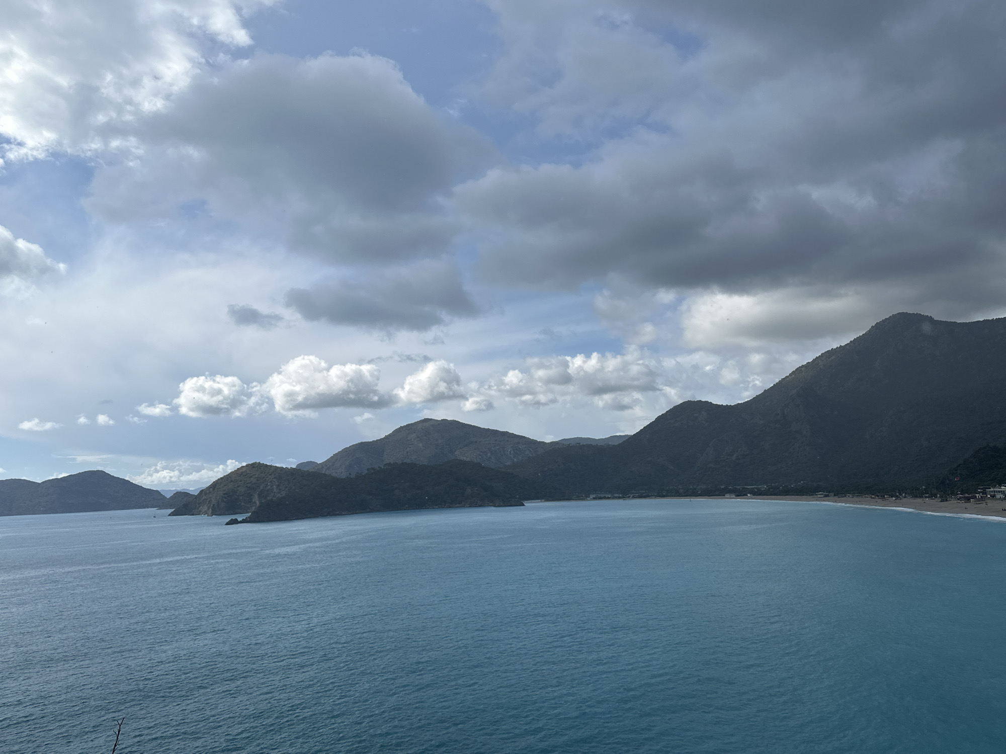 The image shows a scenic view of a bay in Fethiye, Turkey. The turquoise water occupies the majority of the frame. In the background, a series of dark green mountains rise from the shoreline. Several smaller islands are scattered near the left side of the frame. A beach lined with a scattering of buildings stretches along the coast on the right side of the frame. The sky is overcast with a mix of white and gray clouds.