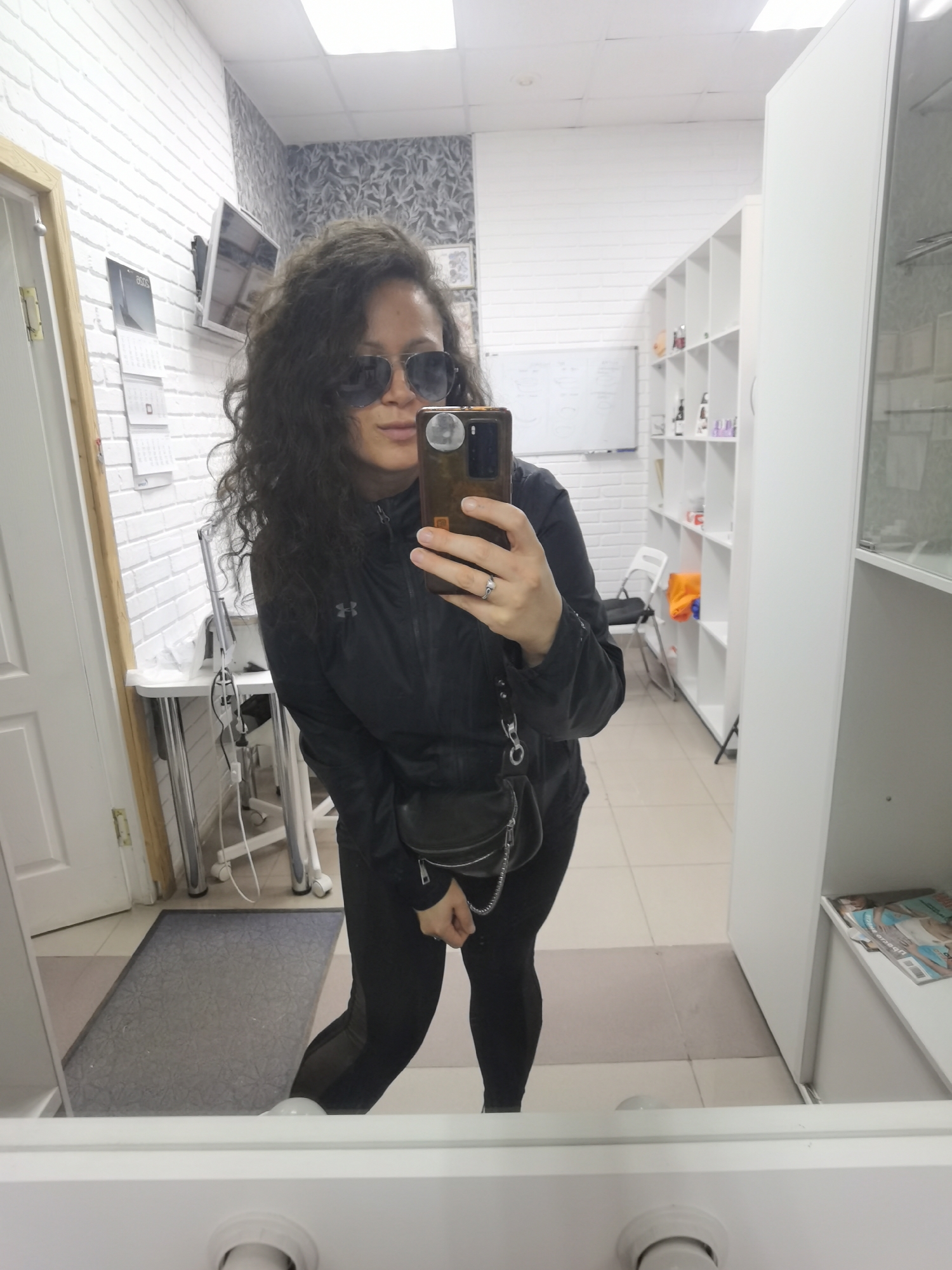 A woman is taking a mirror selfie in what appears to be a salon or treatment room. She has curly dark hair and is wearing sunglasses, a black jacket with a logo, black leggings, and white socks. She is holding a smartphone in her right hand, and a silver ring is visible on her left hand. A small black purse with a chain strap is hanging from her left shoulder. The room has white brick walls, tiled flooring, and shelving units filled with various items. There is a desk with equipment to the left of the woman, and a door is visible in the background. The scene is well-lit by overhead fluorescent lights. No specific details about the city of Chaikovsky, Russia are visible in the image.