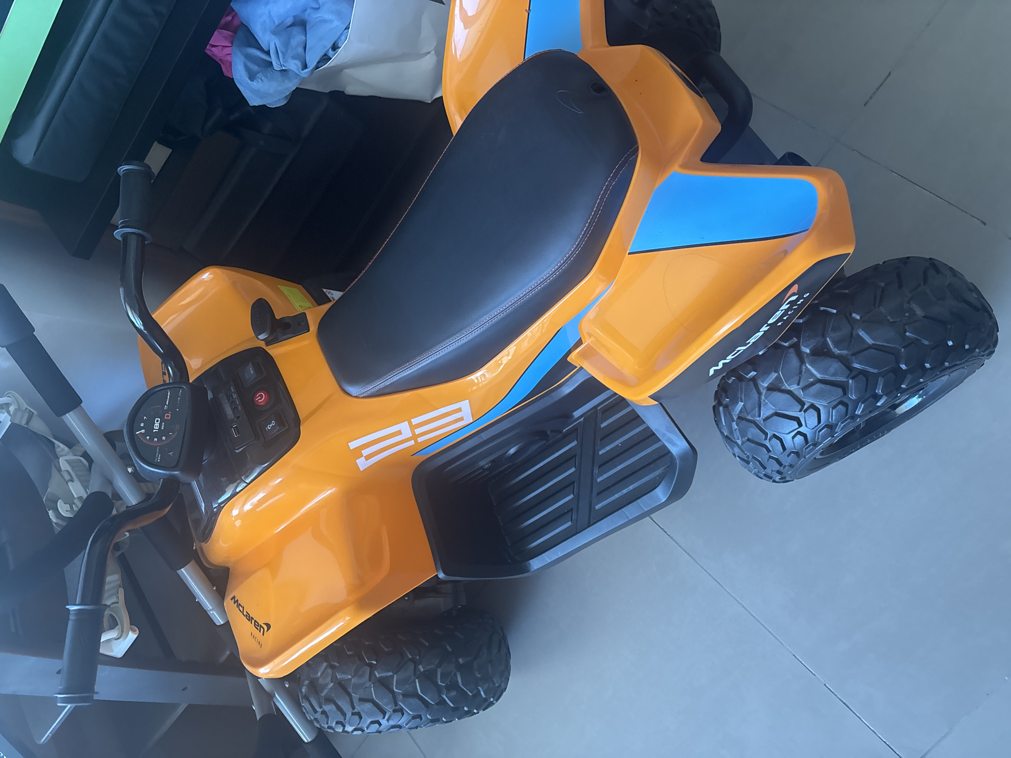 An orange and blue ride-on ATV, possibly a toy, is positioned on a tiled floor. The ATV features black handle grips, a dashboard with a speedometer-like display, and a black seat with orange stitching. Prominent branding on the ATV includes "McLaren Racing" and "EE." In the background, blurred objects suggest an indoor environment, potentially a home, with some laundry visible in a basket to the right. The lighting indicates an indoor setting. The scene is devoid of people and specific Dubai landmarks.