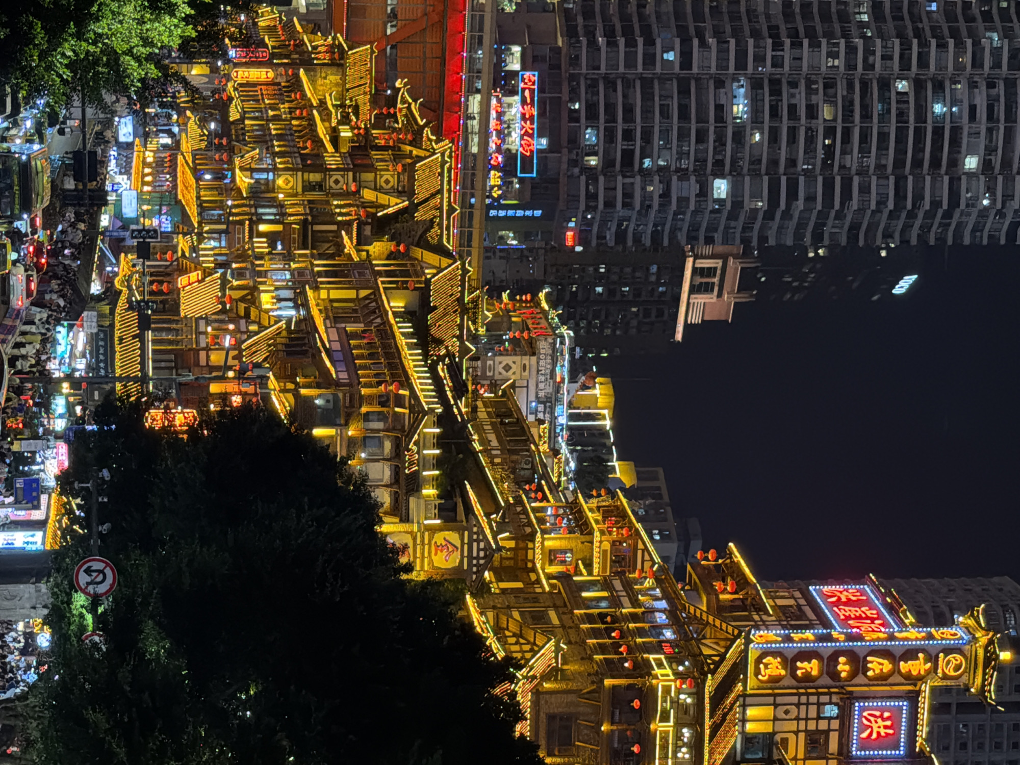 This is a night-time, eye-level, medium shot of the bustling streets and architecture of Yuzhong District, China. The scene is dominated by multi-story traditional Chinese architecture adorned with bright yellow and red lights, intricately detailed with multiple levels of tiered roofs and glowing lanterns. These buildings are stacked on a steep incline, creating a dramatic vertical composition. In the background, a modern, tall skyscraper looms, its windows reflecting faint lights.
The street level is packed with people, suggesting a lively marketplace or entertainment area. Cars, taxis, and buses are visible on the road, indicating active traffic. The atmosphere is vibrant and energetic, highlighted by the colorful neon signs and the dense urban environment. A U-turn traffic sign is visible on the right side of the frame, partially obscured by dense foliage. The overall impression is of a dynamic and visually striking urban landscape at night.