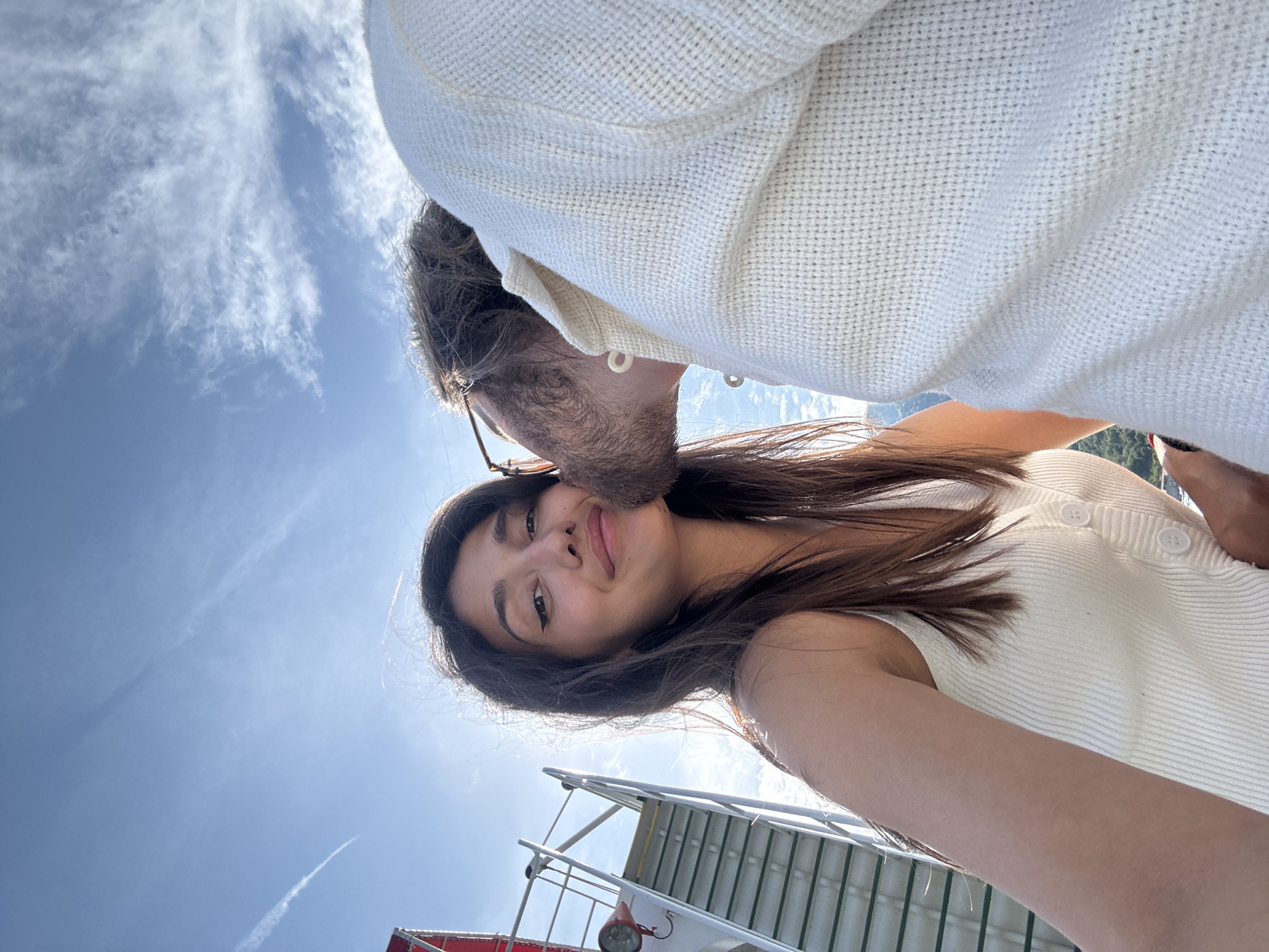 A selfie taken outdoors shows a couple sharing a moment in Prčanj, Montenegro. The woman, in the foreground on the left, has long, dark hair and is smiling as her partner, whose face is partially obscured, leans in to kiss her cheek. She is wearing a white, ribbed tank top with white buttons. Her partner is wearing a light-colored, textured sweater. Behind them, to the left, is a set of stairs with a red railing, suggesting they might be on a boat or a pier. The sky above is blue with wispy white clouds, and the sunlight casts a warm glow on the scene. The backdrop hints at a mountainous landscape, characteristic of the Bay of Kotor.