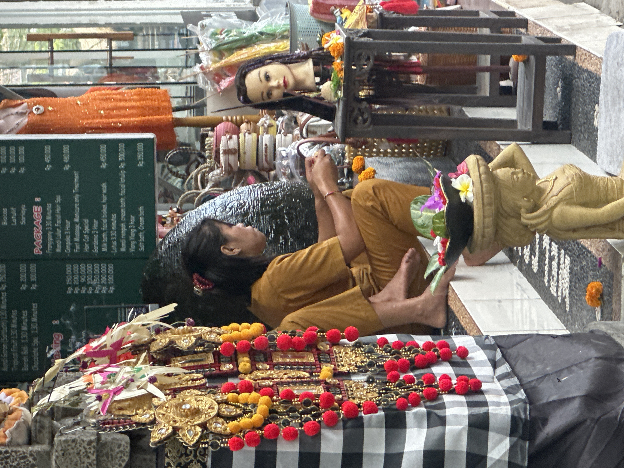 The image depicts an outdoor spa or salon setting in Ubud District, Indonesia, characterized by traditional Balinese decor. In the foreground, a woman wearing an ochre-colored tunic and pants is seated, appearing to be engaged in a task with her hands. To her right, a mannequin head with long braided hair is displayed on a dark wooden stand, adorned with flowers and what appears to be a small tablet. Next to the mannequin, traditional Balinese offerings, likely flower arrangements and incense, are visible.
A prominent feature of the scene is a large, decorative textile with a black and white checkered pattern, hanging on the left side. Adorning this textile are several strings of golden ornaments and red pom-poms, along with other colorful decorations. Behind the woman, a wall made of dark stone or brick is partially visible. In the background, there are shelves stocked with various spa products and decorative items, including woven baskets. An orange, sequined dress is displayed on a mannequin in the middle ground, suggesting a retail component to the establishment. The ground appears to be paved with stones and gravel, with small orange flowers scattered around. The overall atmosphere suggests a tranquil and culturally rich environment.