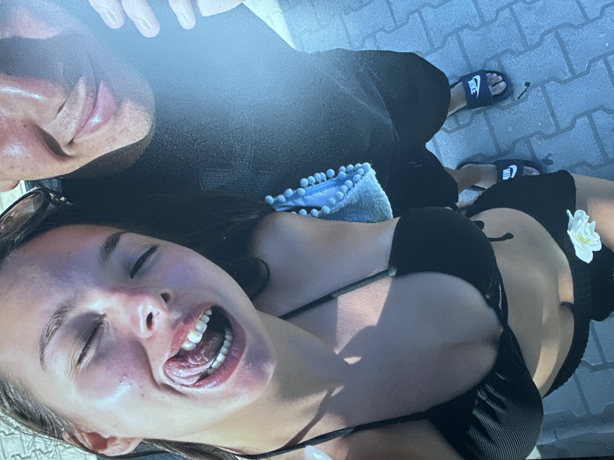 An eye-level, medium-shot photograph captures a young woman with her eyes closed and tongue sticking out, her mouth open in a playful gesture. She is wearing a black bikini top. Beside her, partially visible, is a person wearing a dark t-shirt and black shorts with dark blue Nike slides. The background shows a paved area that appears to be a poolside or patio. The lighting is bright, suggesting an outdoor setting during the day. The woman's skin is glistening, possibly from water or sweat, and she has bright red lipstick on. There is a small white flower attached to the waistband of her bikini bottoms.