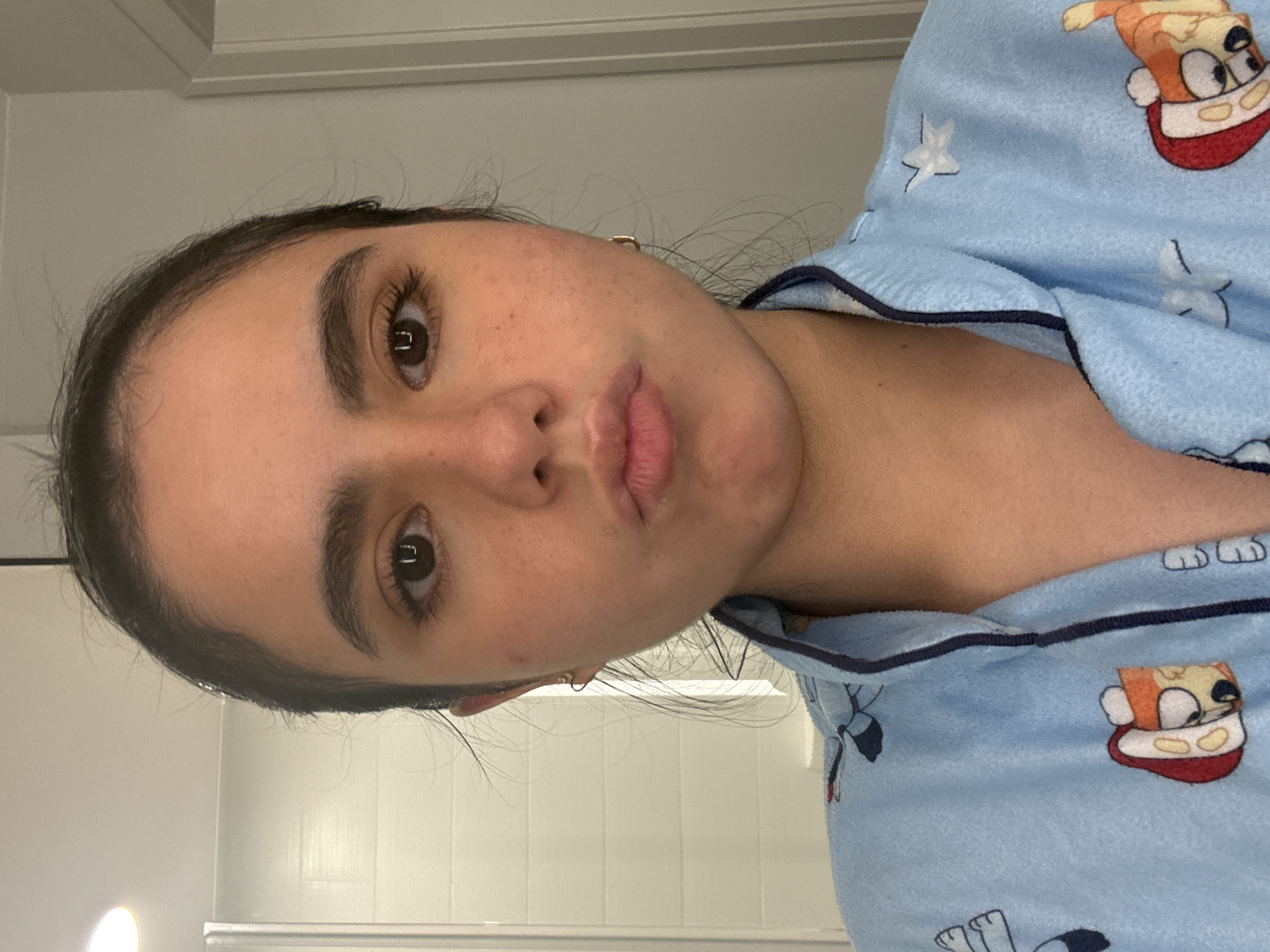 A young woman with dark hair is pictured from the shoulders up. She is wearing a light blue pajama top with an all-over print of cartoon characters. The woman is looking directly at the camera with her lips pursed. Her skin has a smattering of freckles. Subtle lighting suggests the photograph was taken indoors. The background appears to be a bathroom, with a tiled wall and a shower enclosure visible on the left side of the image. The location is not specified as Austin, United States of America.