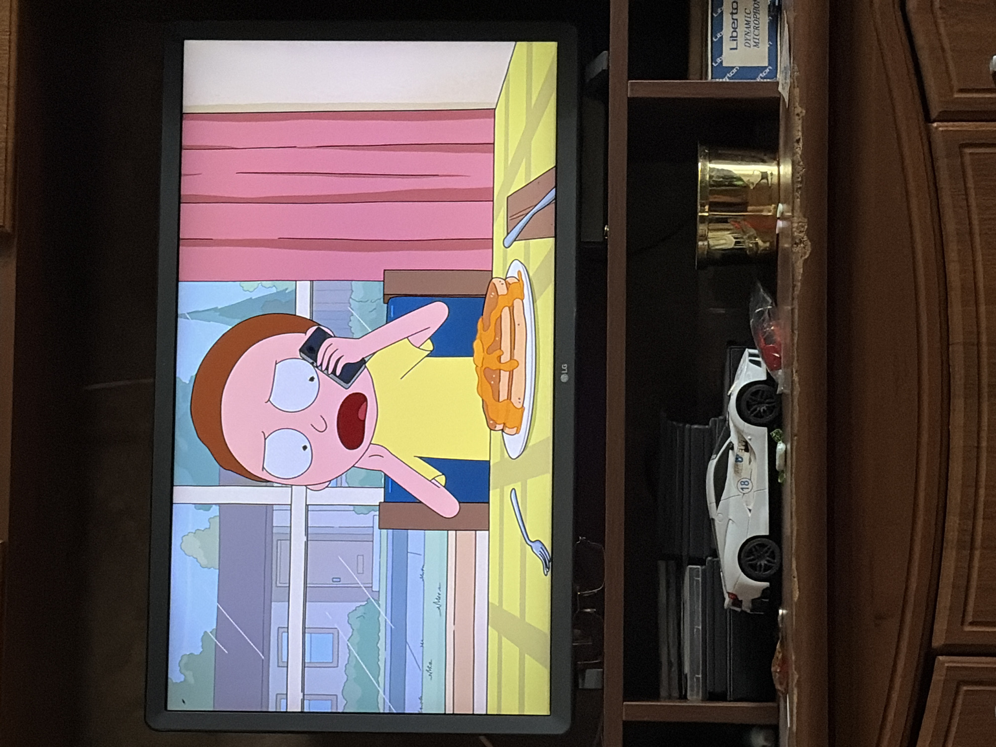 A television screen displays an animated scene of a young boy, identified as Morty, with pink skin and wide, anxious blue eyes, holding a black mobile phone to his ear with his left hand. He wears a yellow t-shirt. Morty sits at a table with a yellow checkered tablecloth. On the table, in front of him, is a white plate piled high with golden-brown pancakes drizzled with orange syrup. A silver fork rests on the tablecloth to the left of the plate. In the background of the animation, a house with a grey roof and light blue siding is visible through a window, with green trees and a bright blue sky. Pink curtains hang to the right of the scene. Below the television, on a wooden shelf, a white toy sports car with black wheels and a circular blue and white decal on its side is positioned to the left. Next to the car, a clear plastic bag contains red objects. To the right of the car is a cylindrical brass object, and further to the right, a blue and white box with text is visible. The television is situated on a dark brown wooden media console.