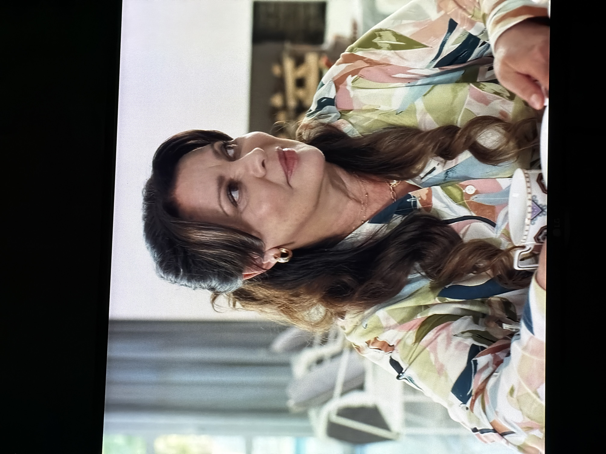 An image shows a medium shot of a woman with brown hair pulled back in a loose ponytail, wearing a patterned blouse and gold hoop earrings. She is looking to her right with a slight smile. She has on a necklace with a delicate gold chain and a pendant. In front of her is a white teacup with a decorative pattern. The background appears to be an interior setting with furniture and a fireplace visible. There is no information or visual cues in the image that would allow for the identification of the location as Semiluki, Russia.