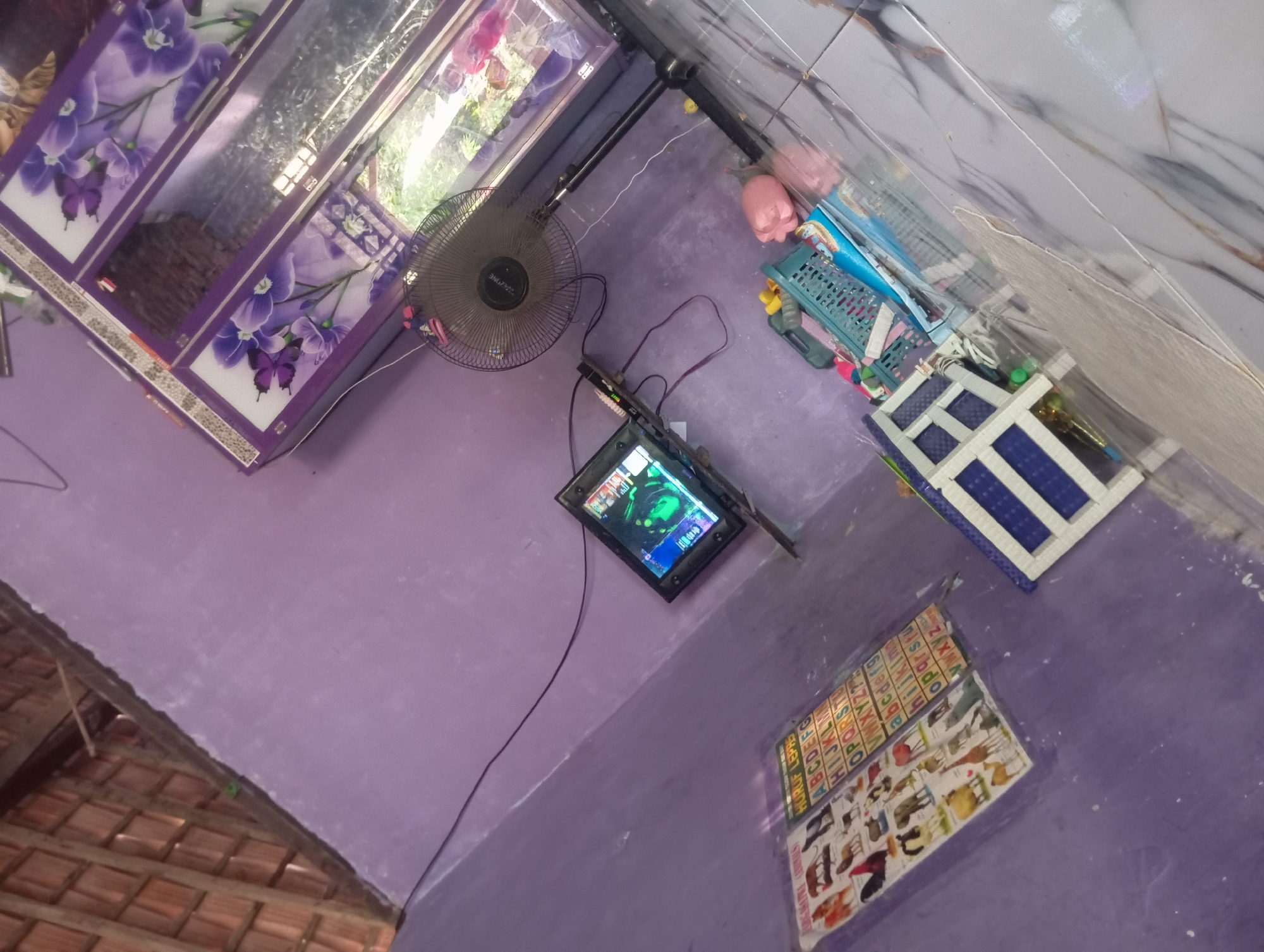 The image shows a corner of a room with purple walls and a tiled floor. In the foreground, there is a collection of colorful toys and a small white shelf unit with blue accents. Behind the toys, a large black standing fan is visible, with a black cord extending from it. To the left of the fan, a small flat-screen television is mounted on a stand, displaying a green and blue image. Below the television, a colorful educational poster featuring animals and letters of the alphabet is displayed on the wall. Further to the left, the ceiling is visible, constructed with wooden beams and terracotta tiles. In the background, a large, ornate cabinet with a floral design is present. The scene appears to be a domestic setting within a residential area.