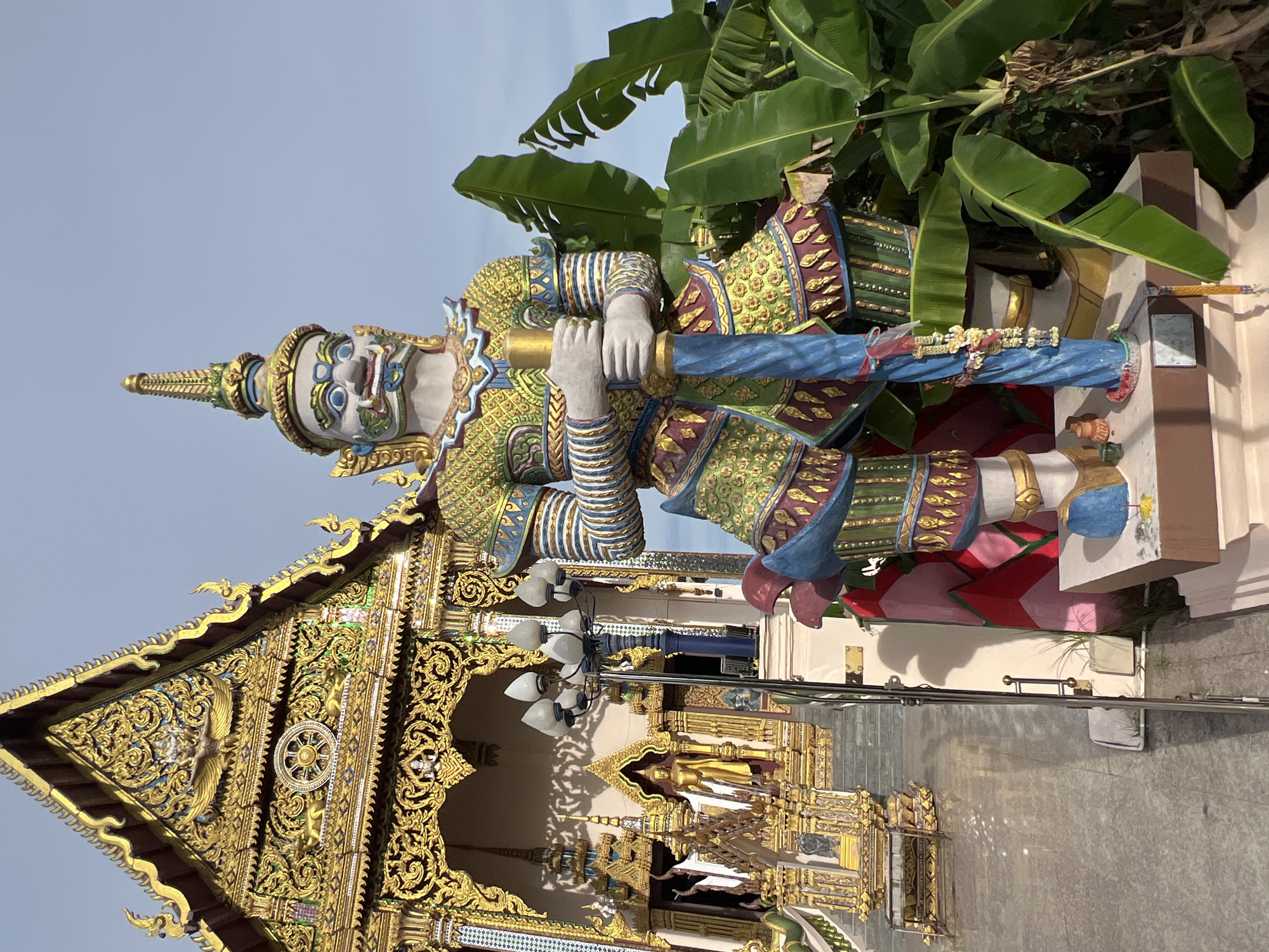This is a photograph of a large, colorful statue of a guardian giant (yaksha) in front of a richly decorated temple building in Baan Plai Laem, Thailand. The statue is depicted standing, holding a long staff or spear. It is adorned with elaborate traditional clothing in shades of gold, green, blue, and red, with detailed patterns and embellishments. The giant has a fierce facial expression with prominent features.
Behind the statue, the temple showcases intricate golden architectural details, including a tiered roof with ornate carvings and decorative finials. A golden Buddha statue is visible within an alcove of the temple. The foreground features a paved walkway, and to the right of the statue, large green banana leaves are prominent. The sky is clear blue, suggesting an outdoor daytime setting. The overall scene conveys a sense of religious and cultural significance.