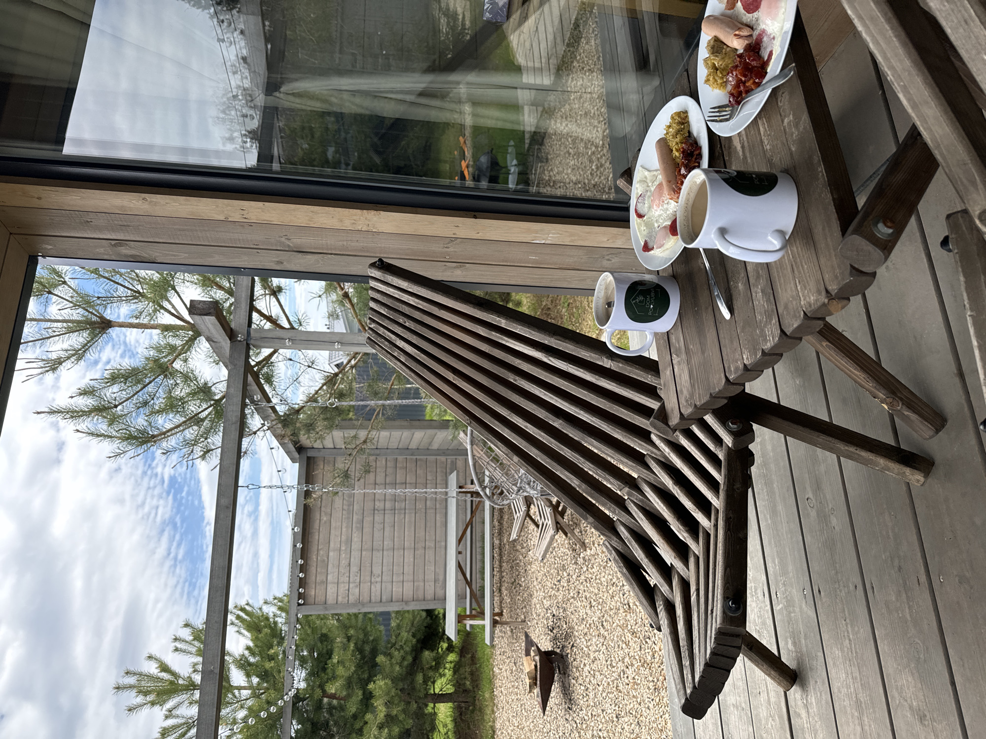 The image depicts a scene on a wooden deck in Литвиново, Russia. Two plates of food, each containing a sausage, potatoes, and berries, along with a fork on one of the plates, are placed on a small wooden table along with two white mugs. The table is situated beside a wooden chair with a slatted back. Through a large window, a view of the outdoors is visible. The outside area includes gravel ground, a wooden picnic table set, a swing, trees, and a clear sky with clouds. A section of the sky is also visible above the deck's structure.