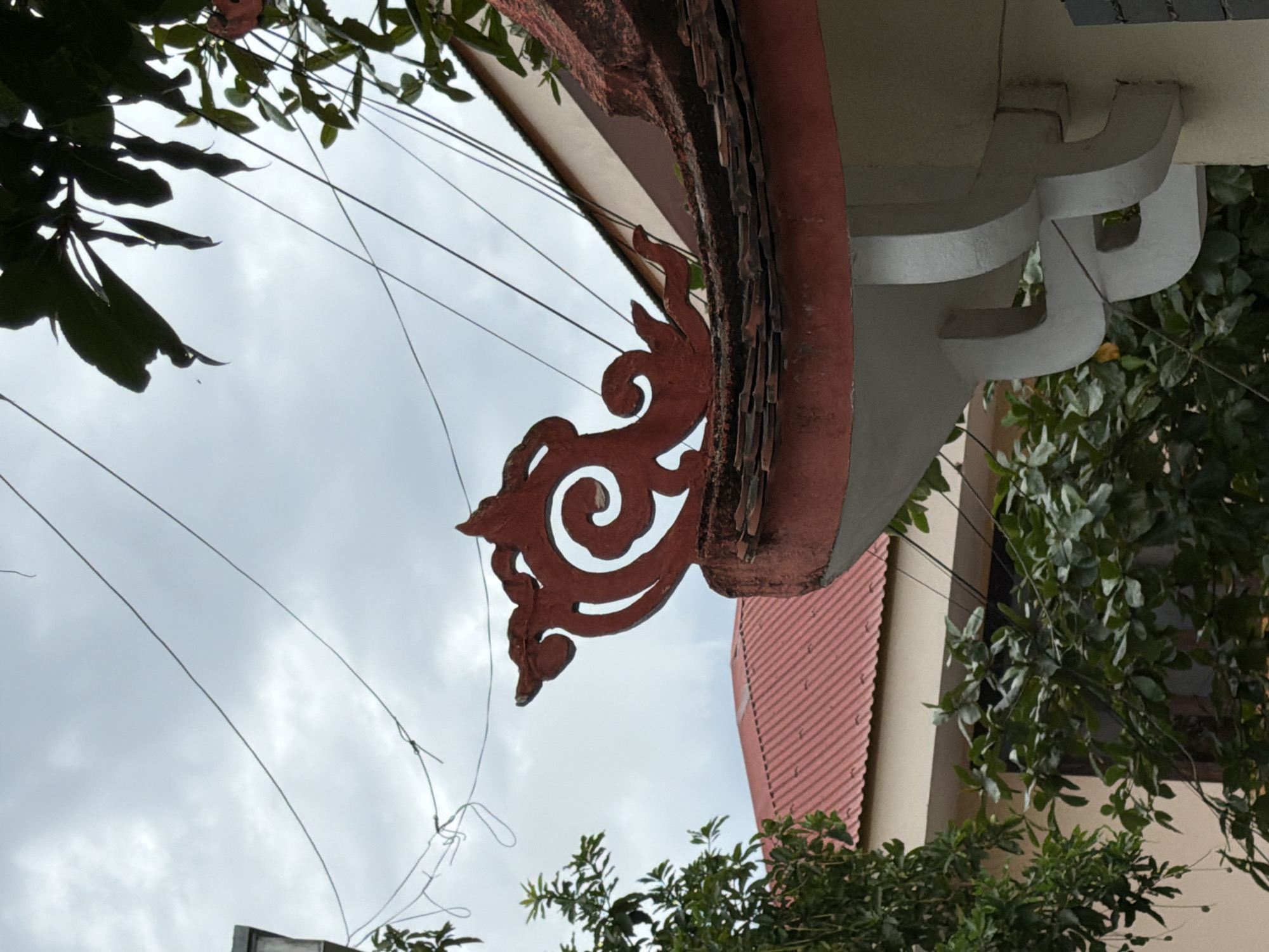 This is a close-up, medium-angle, eye-level shot of the ornate roof trim of a building in Dong Ha Ward, Vietnam. The trim is a vibrant red, intricately carved with swirling patterns, resembling a stylized dragon or phoenix motif. It sits atop a red-tiled roof, partially obscured by green foliage from nearby trees. The background is a cloudy, overcast sky, crisscrossed with power lines. To the left, another building with a red corrugated metal roof is visible. The image focuses on the architectural detail of the trim, suggesting a cultural or traditional building. There are no people or discernible actions depicted. The scene appears to be outdoors.