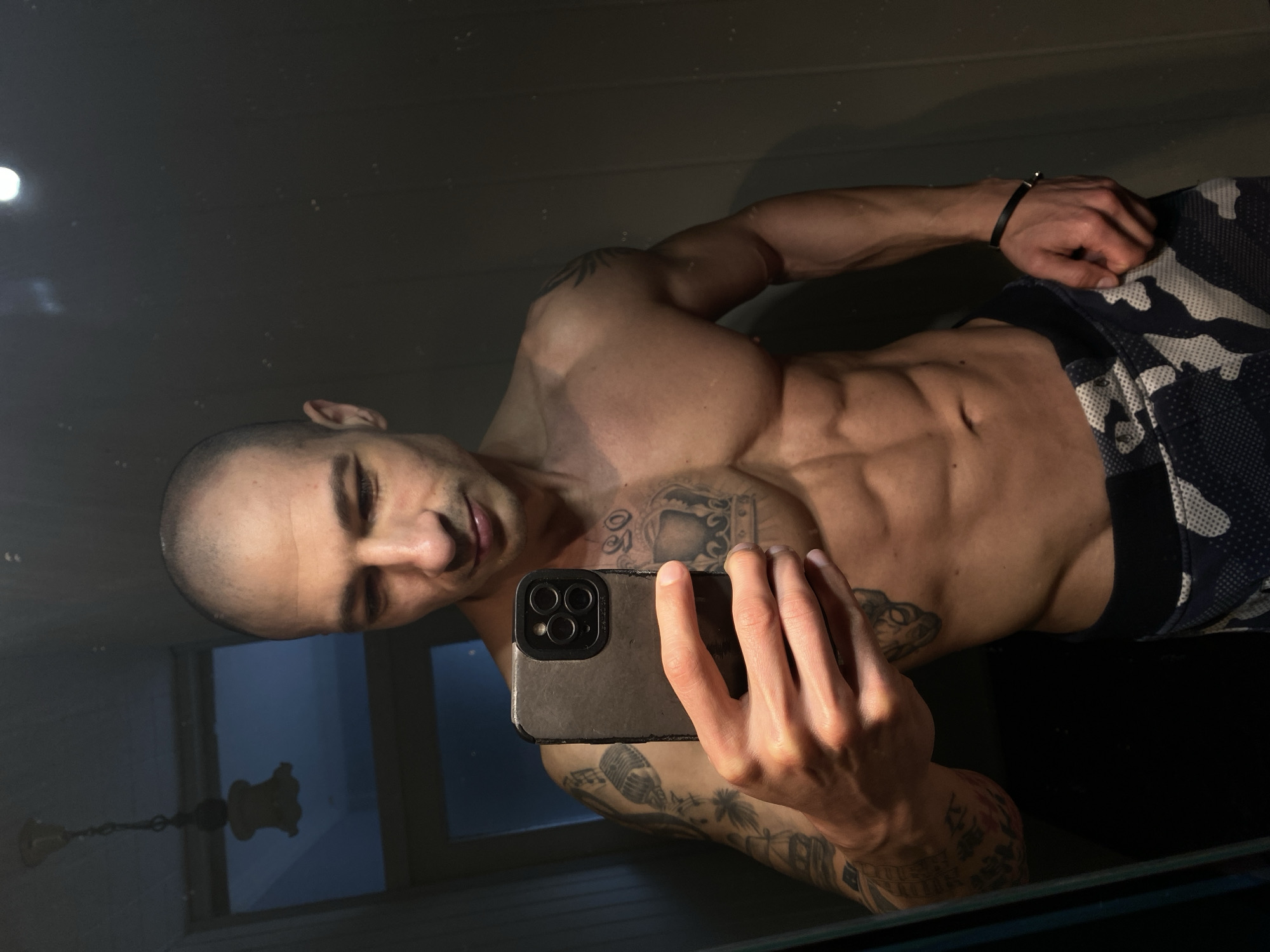 A muscular male individual with a shaved head is depicted in a mirror selfie, positioning his upper torso and part of his lower body. He holds a dark-cased smartphone with a triple camera system in his right hand, angled to capture his reflection. The man's physique displays developed pectorals, deltoids, and abdominal muscles. Visible tattoos include a full sleeve on his left arm featuring a microphone, palm trees, and a ship; a large crown design with the numbers "90" above it on his right pectoral; a tribal-style tattoo on his right shoulder/bicep; and a cat-like face on his right side torso. He wears patterned shorts with a blue, white, and grey camouflage design, and a black leather bracelet with a metallic clasp on his left wrist. The background, visible in the mirror's reflection, reveals a dimly lit indoor setting. To the upper left, a classic-style chandelier with a single light source is reflected. A light blue or grey wall or door is partially visible behind the subject's right side. The mirror surface exhibits small specks or dust. This scene is situated in the city of Pleven, Bulgaria.