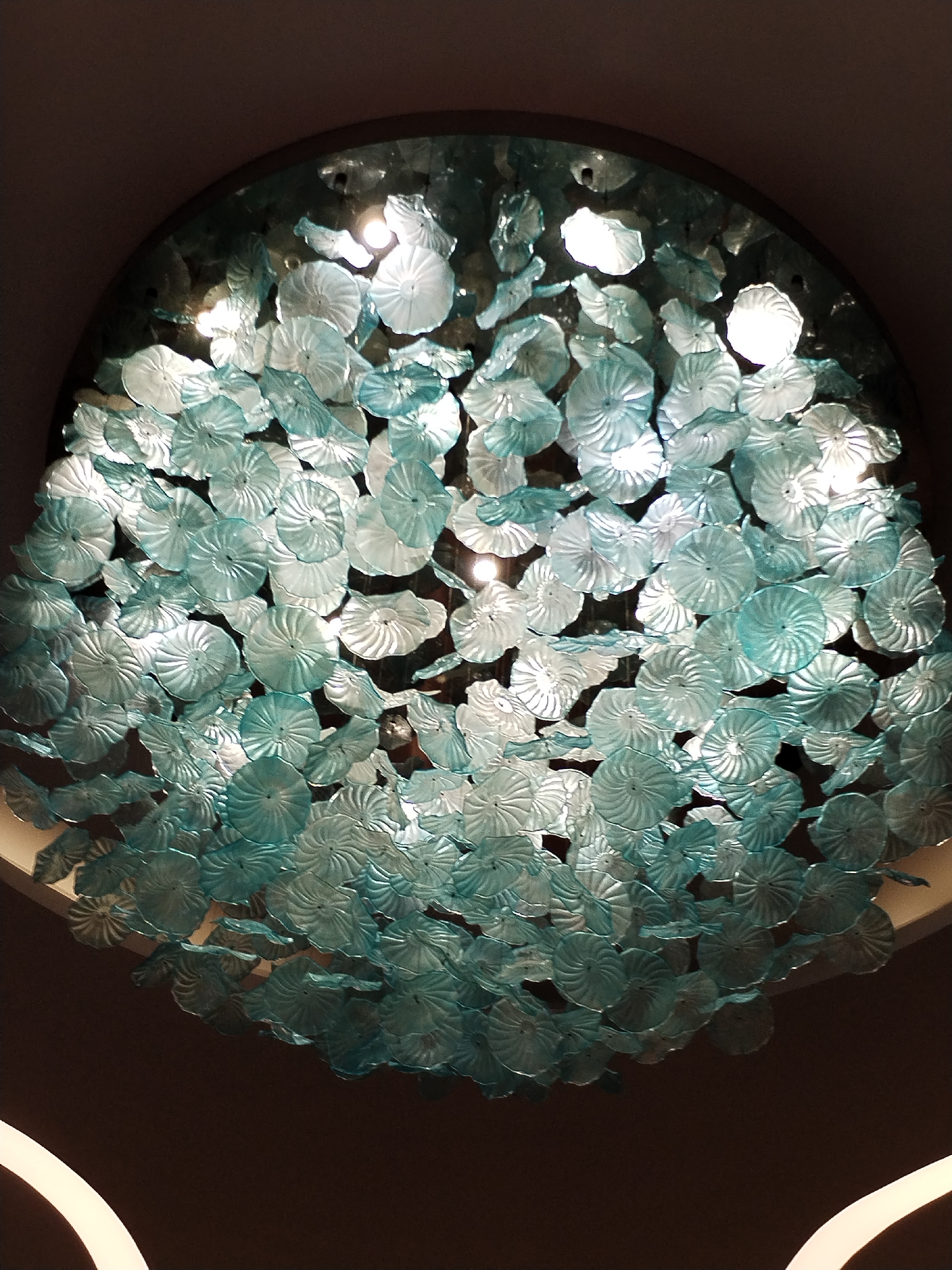 An upward-facing shot reveals a large, rounded chandelier filled with numerous translucent, teal-colored glass elements arranged in a dense, floral-like pattern. These elements are circular with spiral-like patterns etched into their surfaces. Several light sources are embedded within the chandelier, casting bright white light that highlights the translucent material. The chandelier is mounted on a dark ceiling, with the light casting a glow onto the surrounding surface. The presence of an unseen ceiling feature in the lower portion of the image suggests a modern or decorative interior space. While the image provides no immediate clues to the location in Bac Nha Trang Ward, Vietnam, the design and presence of such a chandelier suggest a hotel lobby, restaurant, or upscale commercial space.