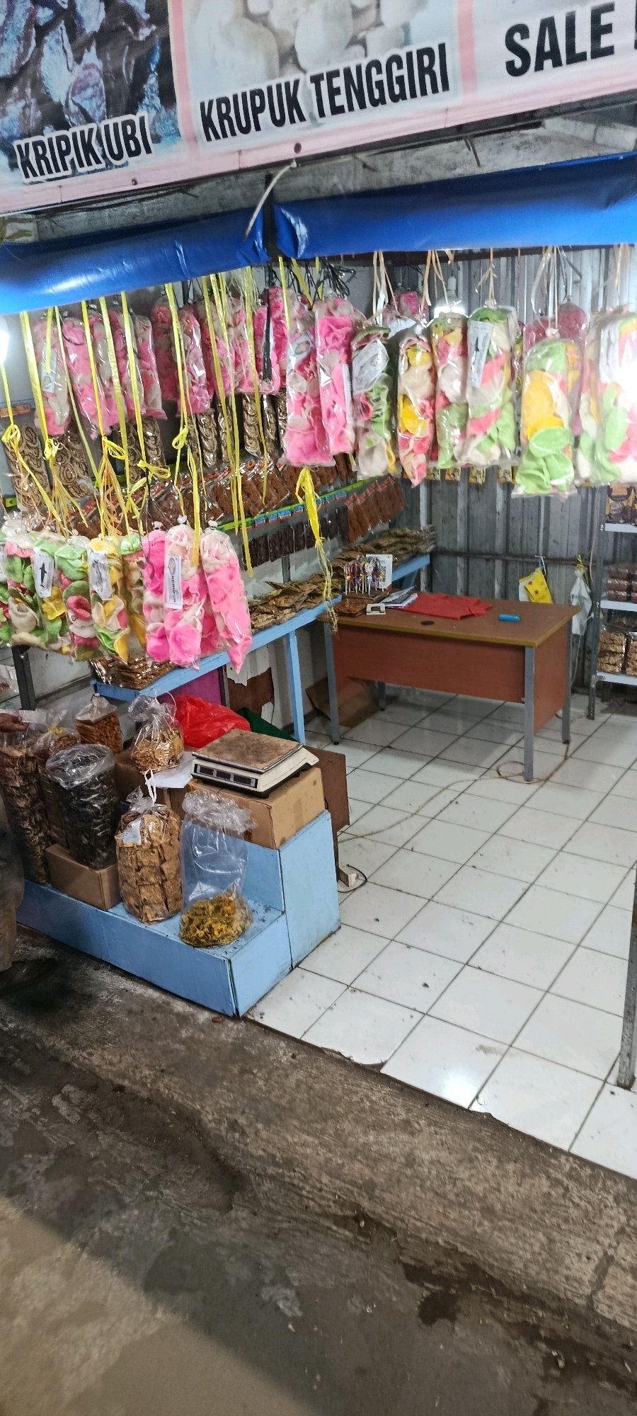 This is a medium-shot, eye-level, outdoor photograph of a small storefront in Megamendung, Indonesia. The storefront is selling various snacks and chips. The products are displayed hanging from the ceiling in clear plastic bags, some with yellow ribbons. There are also bags of snacks on blue tiered shelving units. In the background, there is a desk with a red cloth on it. The floor is tiled. The overall impression is of a humble, local shop.