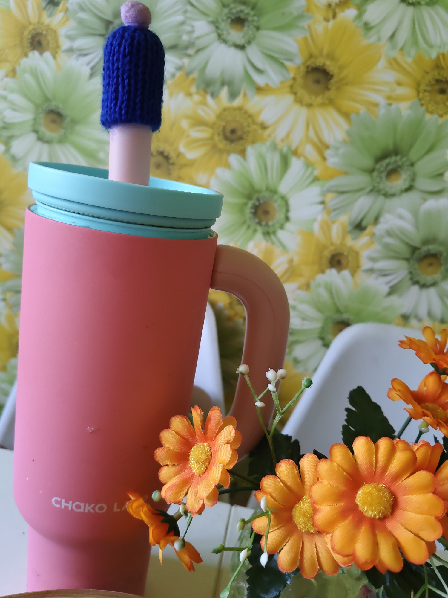 The image shows a close-up of a pink tumbler with a light blue lid, topped with a small knitted blue hat with a pink pompom. The tumbler has the logo "CHAKO LA" printed on its side. In the background, there is a floral wallpaper featuring large daisy-like flowers in shades of yellow, light green, and white. To the right of the tumbler, a bouquet of artificial orange flowers with green leaves and white buds is visible. Two white chair backs are partially visible behind the flowers. The setting appears to be indoors, possibly in a home or a studio, and the overall impression is bright and colorful. There is no indication of the specific location in Rostov-on-Don, Russia.