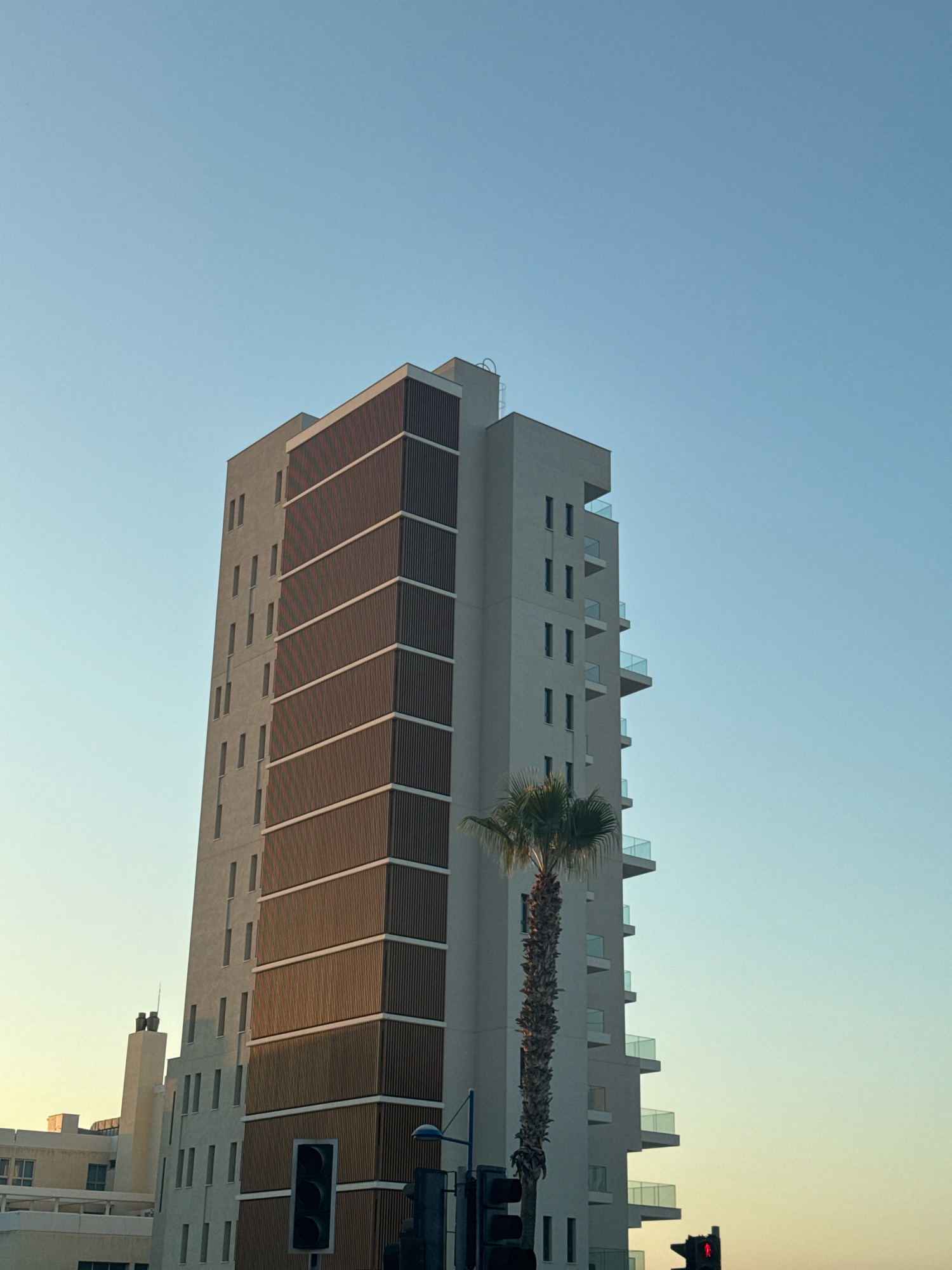 The image depicts a modern, tall, light-colored building with several balconies in Agios Tychona Community, Cyprus. The building features a distinctive section with horizontal wooden slats. A palm tree stands beside the building. In the foreground, a traffic light is visible with a red signal illuminated. The sky is clear and a pale blue, transitioning to yellow near the horizon, suggesting it is either sunrise or sunset.