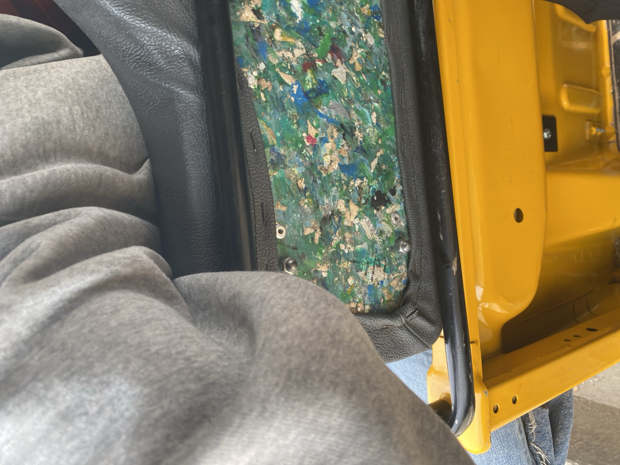 The image shows a close-up view from inside a vehicle, likely a utility vehicle or a truck, painted bright yellow. The foreground is dominated by a person's legs covered in grey sweatpants or similar casual wear, draped over the side of a black leather seat. To the right of the person's legs, the interior of the vehicle's door is visible. This area features a textured black trim bordering a panel made of what appears to be compressed, recycled material, displaying a mosaic of green, blue, white, and red fragments. This panel is secured with visible metal fasteners.
Beyond the door panel, the vibrant yellow painted interior of the vehicle is prominent, revealing structural elements and a mounting point for what might be a component or accessory. The lighting suggests it's daytime, with natural light illuminating the scene. The setting appears to be functional rather than luxurious, hinting at a vehicle used for work or transport. No specific activity is depicted, as the image is a static shot focusing on the interior details. The overall impression is one of a utilitarian vehicle, possibly in a workshop or during a pause in activity. There is no visible text in the image. The location context of Jalingo, Nigeria, suggests this vehicle might be part of local transportation or commercial operations in that region.