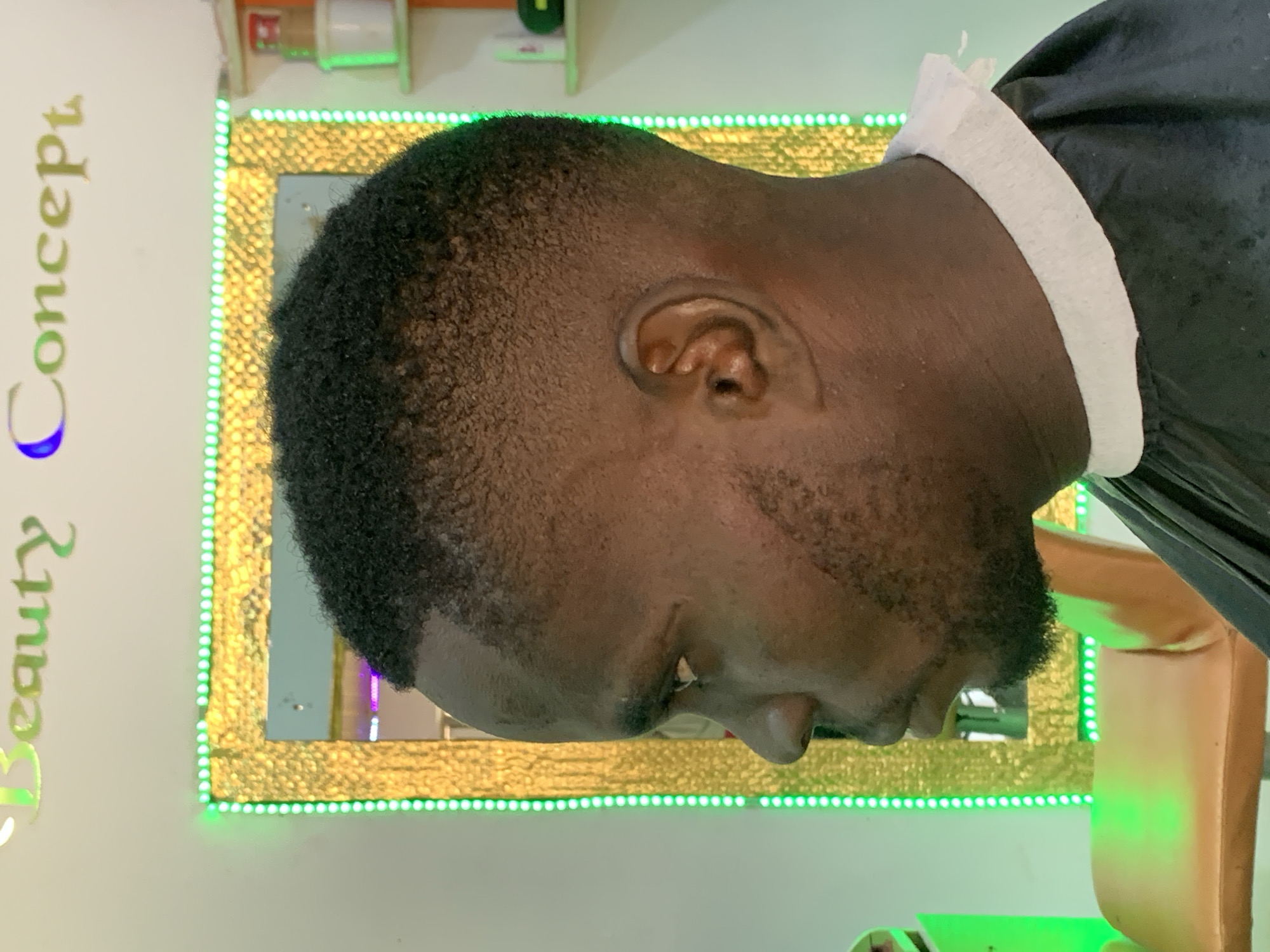 A close-up shot depicts the side profile of a dark-skinned male with a close-cropped haircut and short beard. He is wearing a black barber's cape with a white paper neck strip. The subject is positioned in front of a rectangular mirror with a gold-colored frame illuminated by a string of green LED lights. The words "Beauty Concept" are visible in gold and green lettering on the wall to the left of the mirror, with the final "C" in "Concept" appearing in blue. The location is presumed to be a salon in Ikorodu, Nigeria. The man is either receiving a haircut or is seated in a barber's chair.