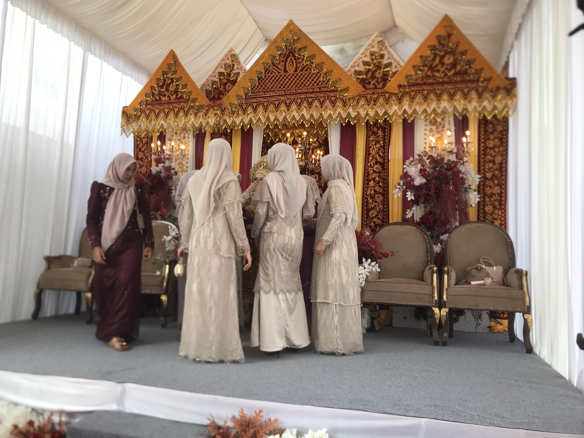 The image captures a formal event, likely a wedding or engagement ceremony, set indoors within a tented structure. A raised stage or dais, covered with grey carpet, serves as the focal point.
On the stage, four women are present. Three women are dressed in elegant, floor-length, light-colored (beige or cream) traditional dresses with intricate lace details and matching hijabs (headscarves). They are seen mostly from the back, gathered around a central figure, possibly the bride, whose face is not fully visible but wears a golden headpiece. The fourth woman, on the left, wears a maroon-colored dress with a light-colored hijab and appears to be walking.
The backdrop behind the women is highly ornate and decorative, featuring a traditional design with multiple peak-shaped golden structures adorned with red and gold patterns. The backdrop is made of rich fabrics in yellow, red, and maroon, embellished with golden accents and beadwork. Two elaborate, upholstered chairs with golden frames are placed on the stage, one holding a light-colored handbag. Floral arrangements with deep red, white, and burgundy flowers, along with illuminated candelabras, add to the celebratory atmosphere.
The event appears to be taking place during the daytime, with ample light illuminating the scene, likely filtered through the white tent walls visible on the left. The attire and decorations suggest a cultural celebration typical of Julimeunasah Teungoh, Indonesia. No visible text is present in the image.