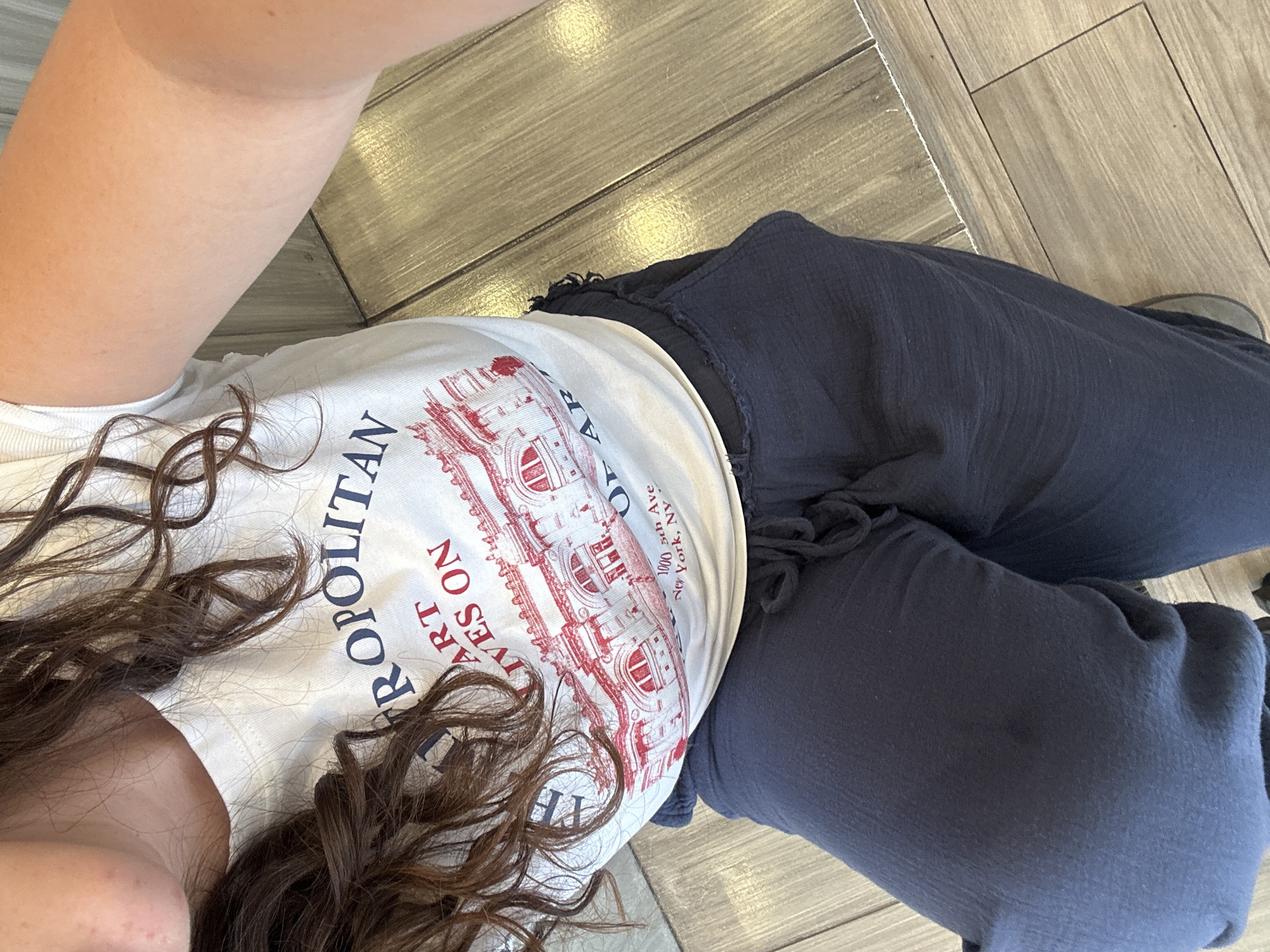A person is lying on a floor with wood-like tiles, wearing a white t-shirt with a graphic depicting the Metropolitan Museum of Art, along with the text "THE METROPOLITAN ART LIVES ON 1000 5th Ave New York, NY," and dark blue, loose-fitting pants. Part of the person's face and long, curly brown hair are visible.
