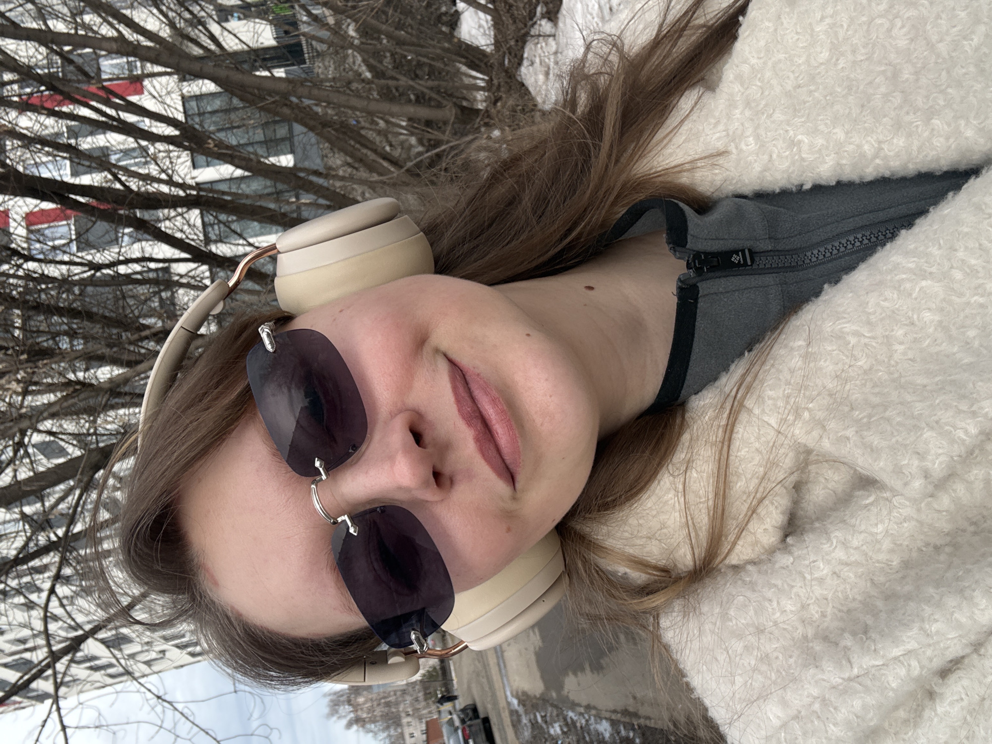 A woman is wearing sunglasses and headphones, and a white fuzzy jacket or coat, outdoors. The background appears to include a city building and leafless tree branches. The location could be in Izhevsk, Russia, given the request, though specific landmarks are not clearly visible.