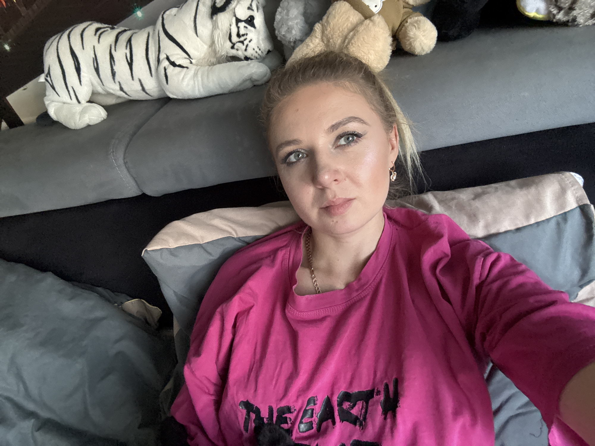 In an indoor setting, a young woman with blonde hair pulled back in a ponytail takes a selfie. She is lying down, with a plush white tiger with black stripes to her left. Other stuffed animals, including a tan bear and a gray stuffed animal, are visible behind her. The woman is wearing a bright pink long-sleeved shirt with black lettering that reads "THE EARTH IS OUR HOME". She has makeup on, including eyeshadow and eyeliner, and is wearing a gold necklace and small gold earrings. The background consists of a sofa and pillows. The lighting appears to be natural, coming from the front.