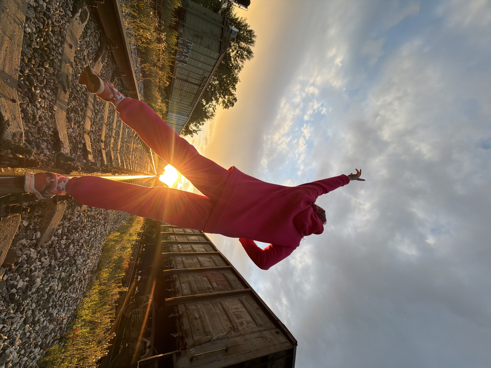 A person, wearing a pink hooded sweatshirt and matching pants, is captured in a dynamic pose on a set of train tracks during sunset. The individual's body is angled diagonally across the frame, with one leg extended forward and the other bent back, creating a sense of movement and freedom. Their arms are outstretched, one reaching towards the sky.
The setting is an outdoor train yard, with the tracks running diagonally through the image. Several weathered, dark-colored freight train cars are visible, suggesting an industrial or abandoned location. The ground beside the tracks is covered in gravel, with patches of green grass and vegetation growing along the edges.
The time of day is clearly sunset, indicated by the warm, golden light that bathes the scene and creates strong lens flare from the sun. The sky is filled with clouds, some illuminated by the sunset's glow, creating a dramatic backdrop. The overall mood is one of exhilaration and adventure, amplified by the person's energetic pose and the striking natural light.
There is no visible text in the image.