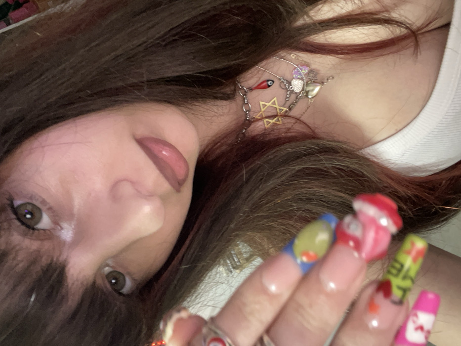 A woman's face is angled toward the camera. She has brown hair with red highlights, brown eyes, and glossy lips. She wears earrings with a Star of David and other charms. Her nails are long and painted with various designs, including food-themed decorations like a mushroom and other colorful images. The background is blurred.