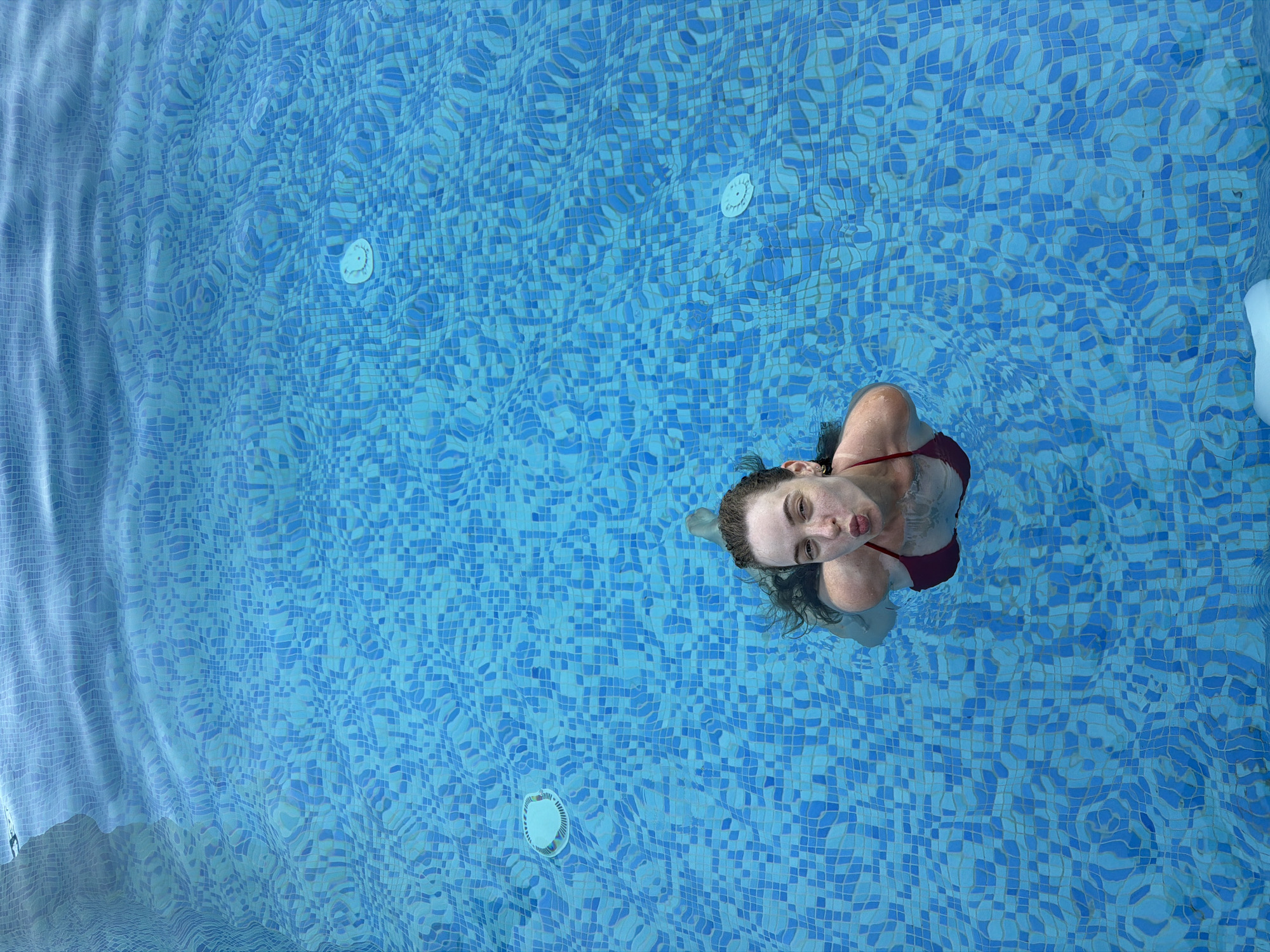 A woman in a red bikini swims in a blue tiled swimming pool. She is looking up at the camera with her lips pursed. The water is rippled and clear, and the sunlight is reflecting off the surface. There are two white circular jets visible in the pool. The scene appears to be located at a resort or private residence within Agios Tychona Community, Cyprus.