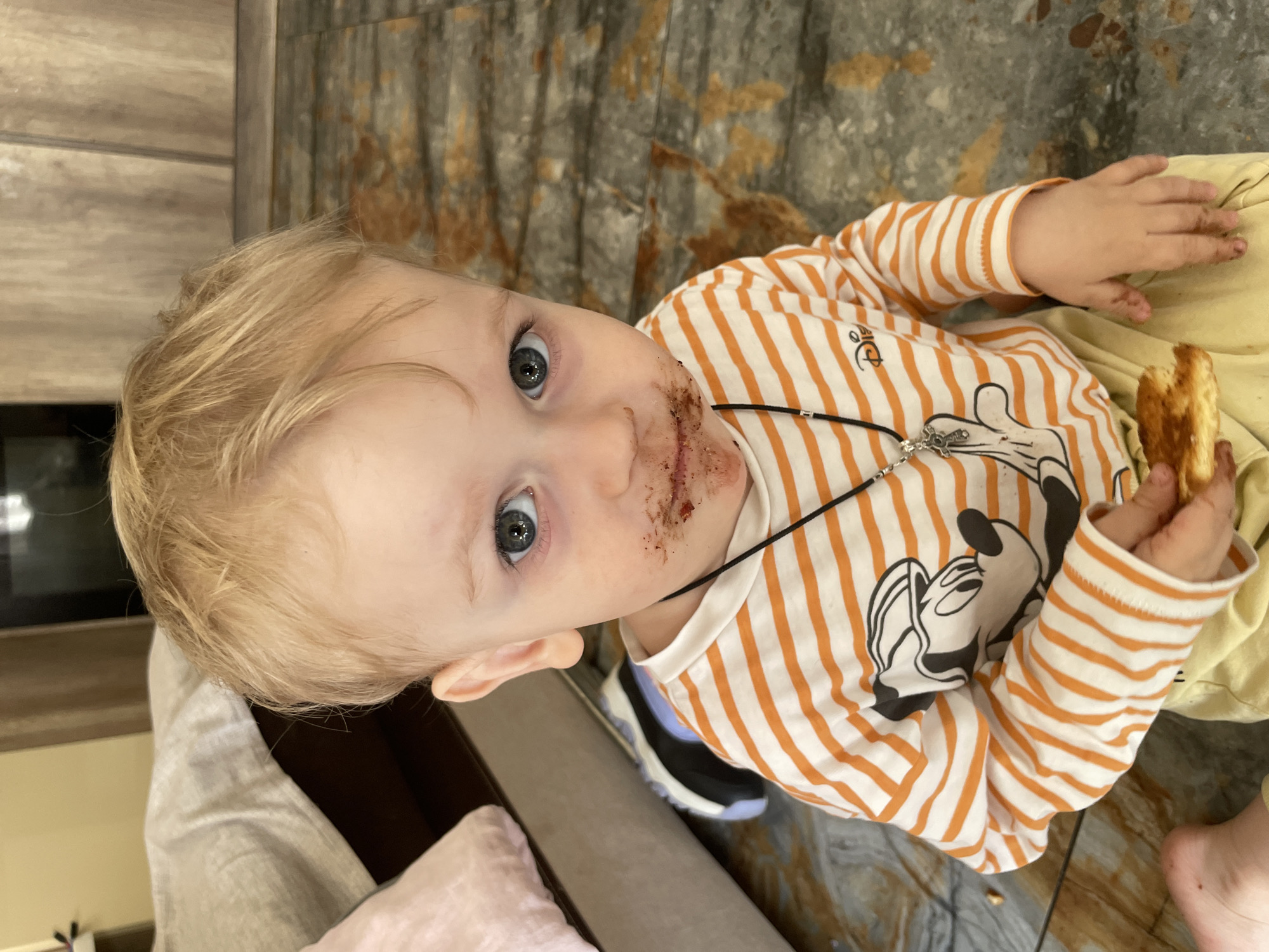 A young child with fair hair and blue eyes is looking upwards, appearing to be the focal point of the image. The child has smudges of what appears to be chocolate or a dark food substance around their mouth. The child is wearing a long-sleeved shirt with orange and white stripes, featuring a Mickey Mouse design. Around the child's neck, there is a thin black cord or necklace with a silver-colored charm hanging from it. The child is holding what seems to be a piece of toasted bread or a similar baked good in their right hand.
The child appears to be sitting or lying on a surface that includes beige fabric, possibly part of a couch or blanket. Partially visible in the background is a floor with a mottled pattern in shades of gray and brown. The setting gives the impression of being indoors, possibly in a residence. The city and location are unknown.