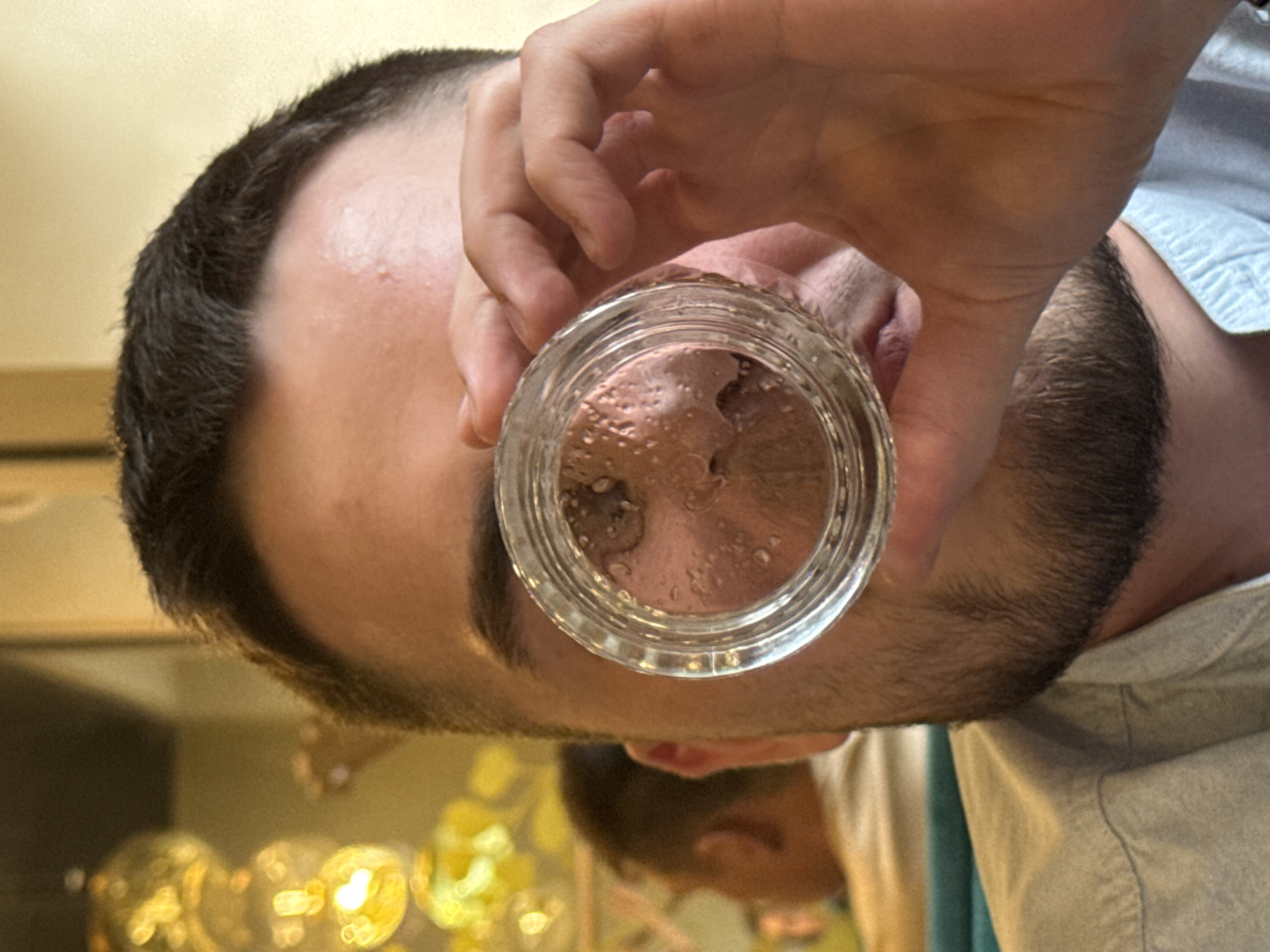 A person is holding a glass jar containing an unidentified brown substance in front of their face. The jar is circular with ribbed sides, and the contents appear viscous or lumpy. The person is light-skinned with dark hair and a beard. The background is blurred, but suggests an indoor setting with possible decorative objects or lights in the background. There is insufficient information in the image to determine the city of Perm, Russia or further details.