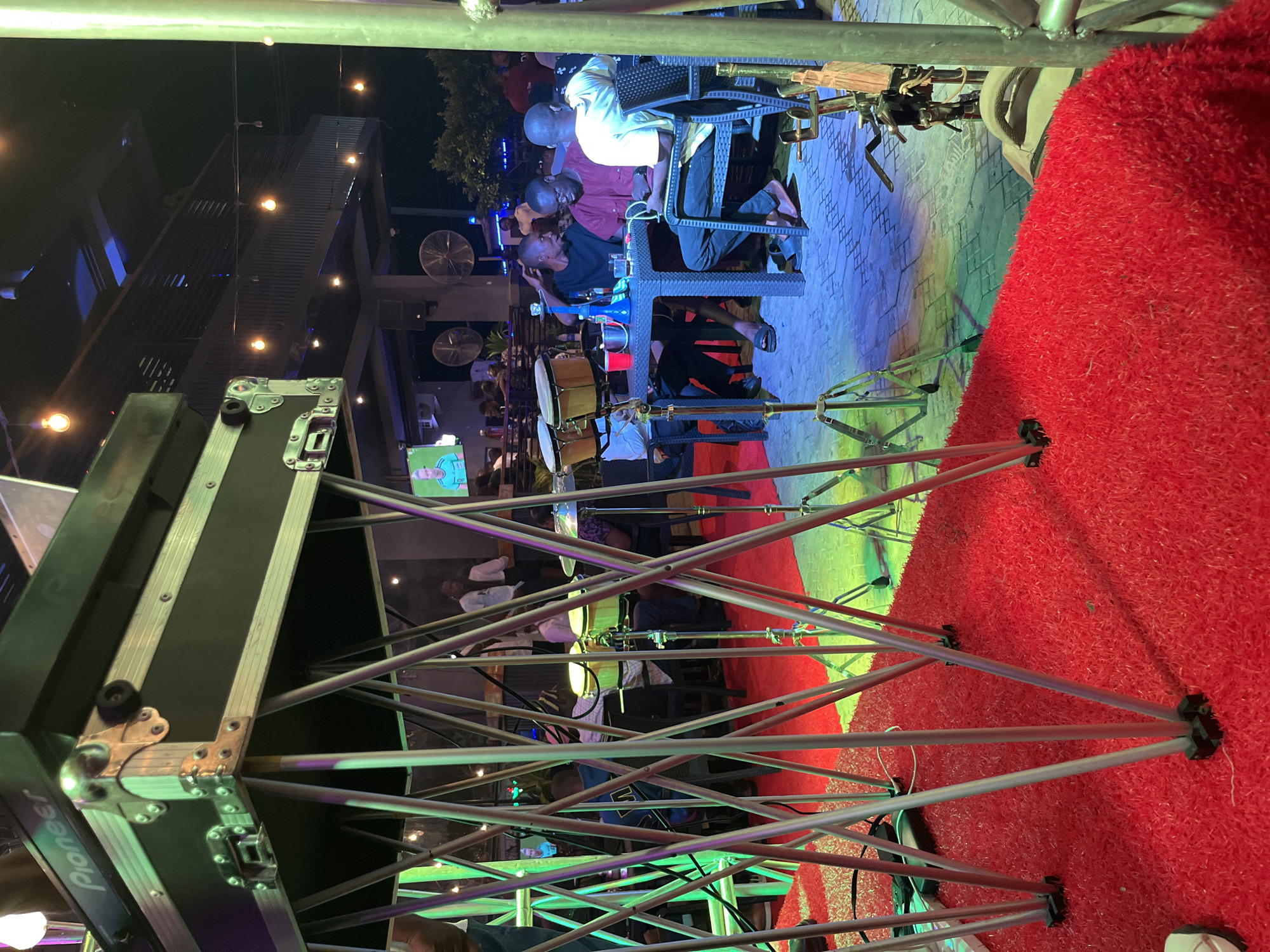 This media file depicts an outdoor evening event, possibly a live music performance. The scene is lively with a crowd of people seated at tables, suggesting a restaurant or bar setting. A drum set is visible, indicating that a musical act is either performing or about to perform.
In the foreground, a large black case with "Pioneer" visible on it is prominent, likely containing audio equipment. The ground is covered in a vibrant red carpet or artificial turf, adding a festive touch. In the background, there are elevated walkways and seating areas, illuminated by various lights that cast colorful reflections on the ground, creating a dynamic atmosphere.
Several individuals are seated at tables, some engaged in conversation. The lighting suggests it is nighttime. The overall mood appears relaxed and enjoyable, typical of a social gathering with entertainment. The image is oriented vertically, with the top of the scene appearing on the left.