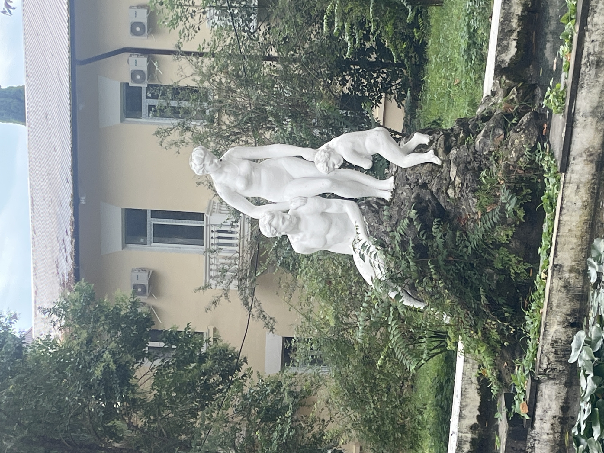 A white marble sculpture depicts a family group in a garden setting. A male figure, seated, is positioned to the left, while a standing female figure is at the center, holding the hand of a small child who is to the right and slightly in front of her. The sculpture is placed on a rock formation overgrown with green foliage, located near a concrete pond with lily pads. In the background, a pale yellow building with a grey, sloped roof and white-trimmed windows is visible. Two air conditioning units are mounted on the exterior wall of the building. The scene is likely located in Sokhumi, Abkhazia, given the architectural style and the lush vegetation, typical of the region's climate. The lighting suggests an overcast day.
