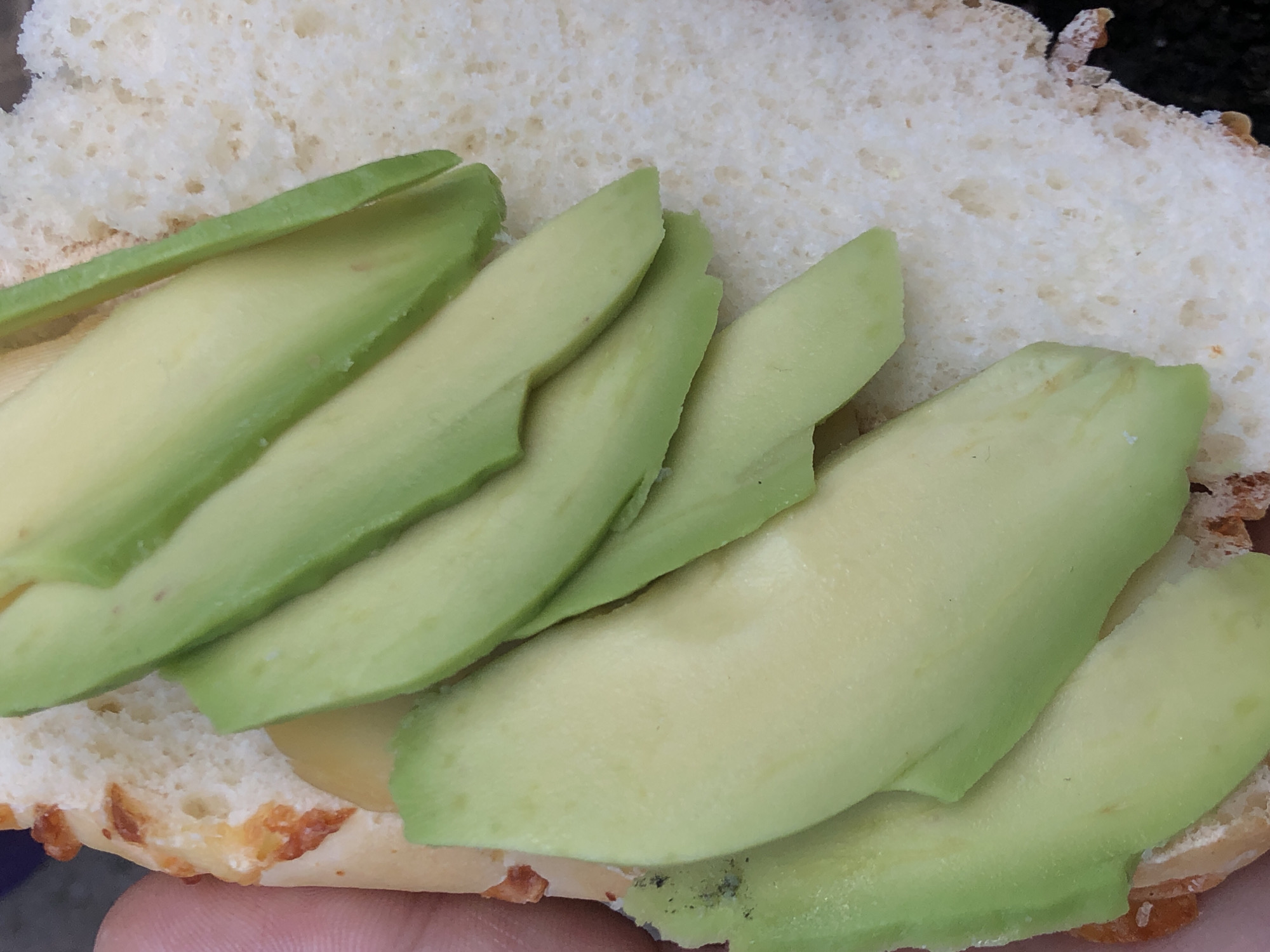 A close-up, top-down shot captures a food item consisting of sliced avocado on a piece of white bread. The main subject is a layered arrangement of thin, bright green to light green avocado slices, covering a significant portion of the bread. The avocado appears fresh and ripe, with some slices showing slight browning or bruising on their edges.
The bread underneath is white with a soft, porous texture, resembling a baguette or a type of roll. Along the bottom edge and partially on the sides, the bread features golden-brown, irregular patches, possibly indicating baked cheese or a crispy crust. A human finger is visible at the very bottom of the frame, holding the food item, suggesting it is a handheld snack or meal. The lighting is bright and even, highlighting the textures of the avocado and bread. No specific location or time of day can be determined from the image. There is no visible text.