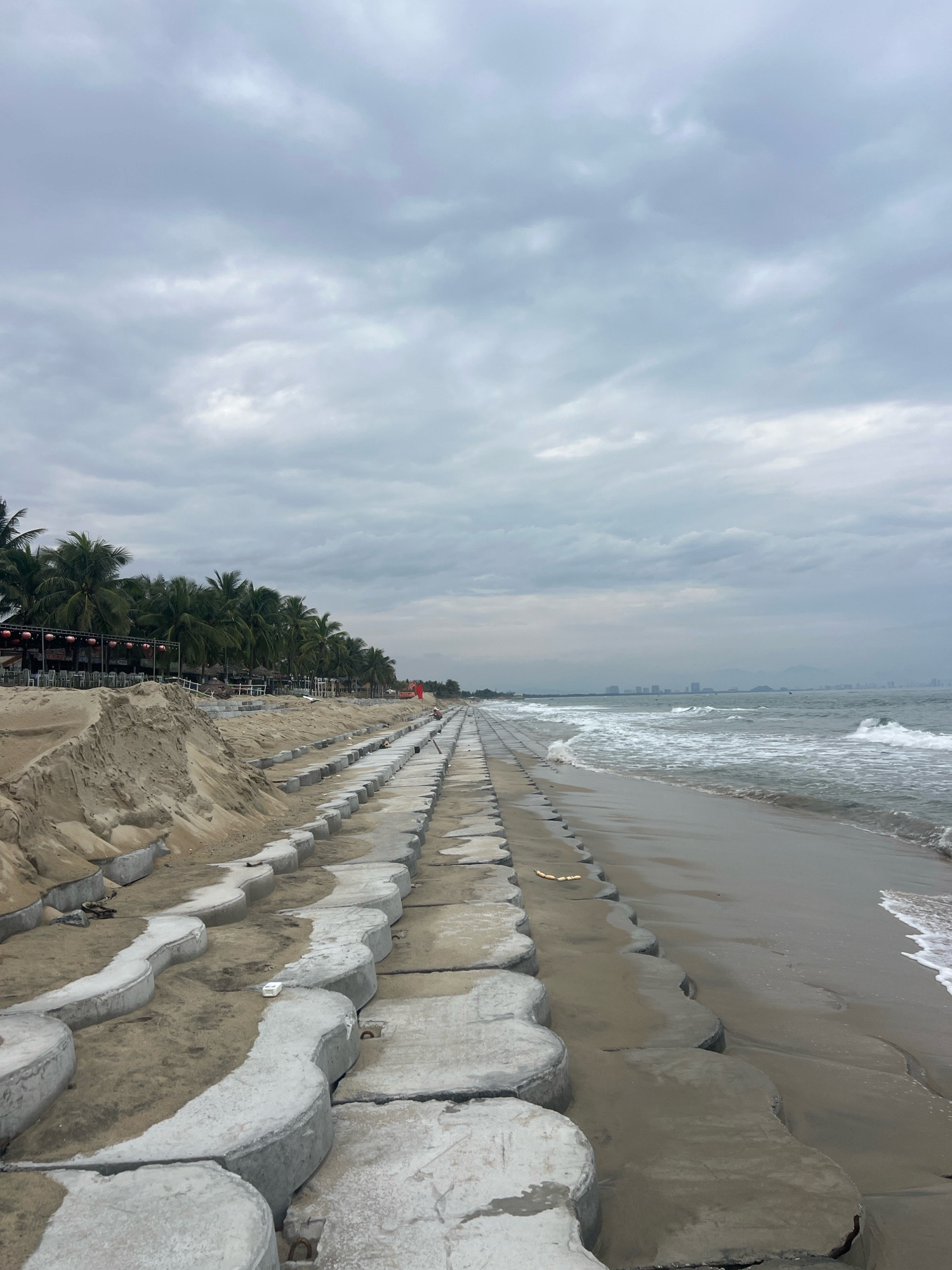 This image depicts a beach in Hoi An, Vietnam, featuring a shoreline reinforced with interlocking concrete blocks. The overcast sky suggests a cloudy or possibly stormy day. Along the left side of the image, a sand dune has formed, contrasting with the orderly placement of the concrete structures. Behind the sand dune, a row of palm trees marks the edge of the beach, with some trees showing signs of wear or damage. Further back, there are indications of a developed area, with what appear to be restaurant tables and chairs visible under a canopy. A red flag, possibly for safety or signaling, is planted near the palm trees. The sea on the right is moderately rough, with waves gently breaking on the shore. In the distance, a hazy cityscape with buildings and hills is visible. The concrete blocks extend along the entire visible length of the beach, suggesting an ongoing or recent construction project to combat coastal erosion. There are no people clearly visible in the frame, though a distant figure may be present in the far right middle ground.