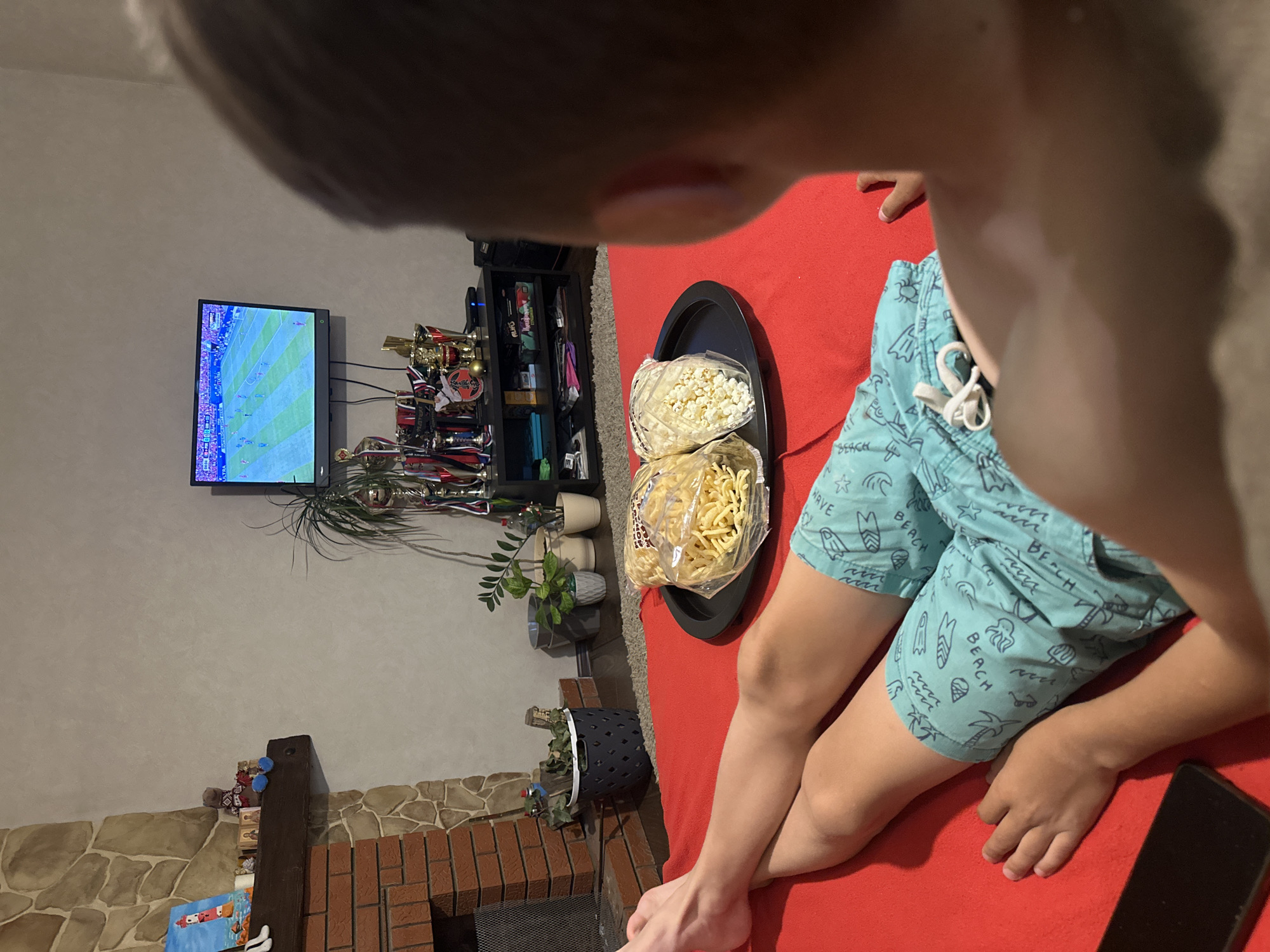 A close-up, overhead shot shows a young person sitting on a bright red surface, possibly a couch or bed. They are wearing light blue swim trunks with a pattern of beach-themed images, including surfboards, palm trees, ice cream cones, and the word "BEACH" repeated. The person's legs and arms are visible, with their right hand resting on their thigh.
In front of the person, on a dark tray, are two bags of snack food. One bag is filled with what appears to be popcorn, and the other contains a yellow, string-like snack.
To the left of the person and behind them, a television screen displays a soccer game. Below the TV, a shelf unit is filled with various items, including trophies, medals, and potted plants. A stone fireplace is visible in the lower left corner of the frame, with a dark wooden beam above it.
The lighting suggests it might be daytime or that the room is well-lit. The presence of swim trunks, the beach-themed pattern, and the relaxed posture of the person suggest a casual, leisure activity, possibly a warm day or a vacation setting. The soccer game on TV could indicate an interest in sports. The overall impression is one of a comfortable, indoor setting where someone is enjoying snacks while watching television.