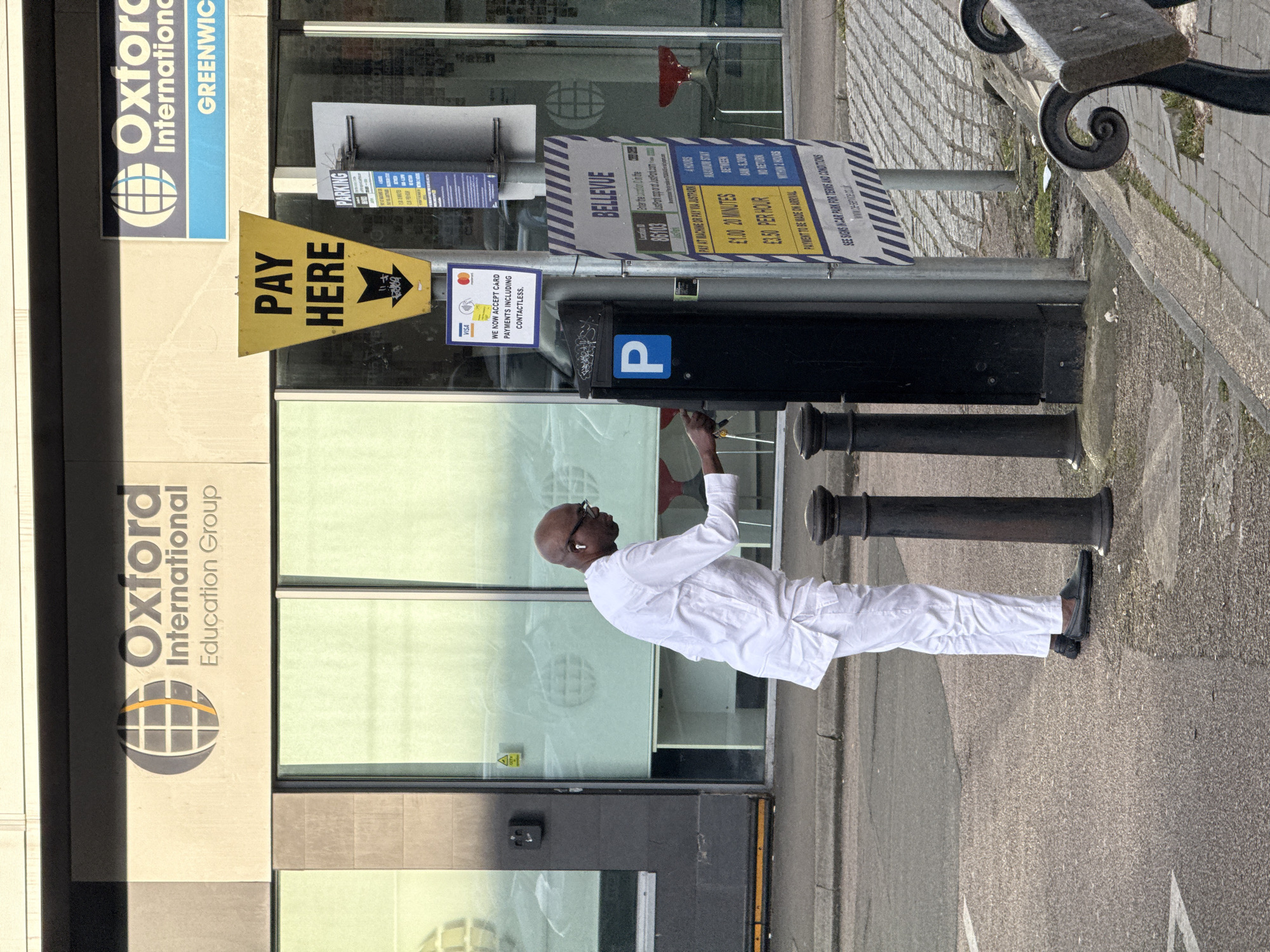 The image captures a man in a white suit at a parking meter on a London street. He's paying using contactless payment. Behind him, the "Oxford International Education Group" and "Oxford International Greenwich" signage are visible. A "PAY HERE" sign and parking rate information ("BELLEVUE: £1.00 20 MINUTES, £3.50 PER HOUR") are displayed on the meter. The location appears to be an urban area with brick paving.